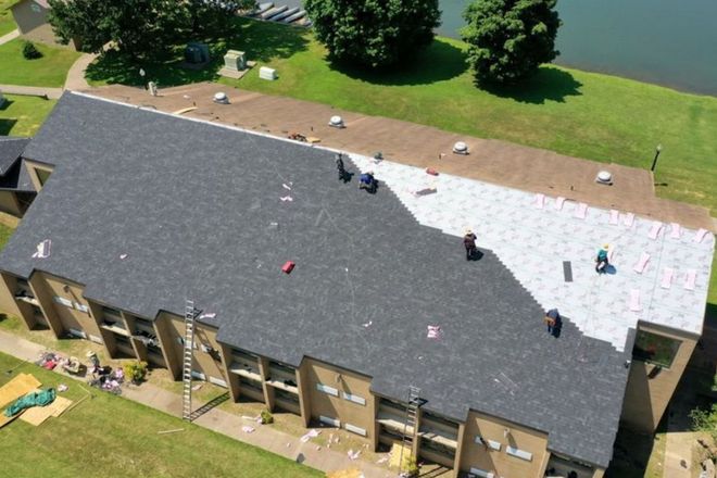 Roofers working on a building with a newly installed dark gray shingle roof, with a light colored section, near water.