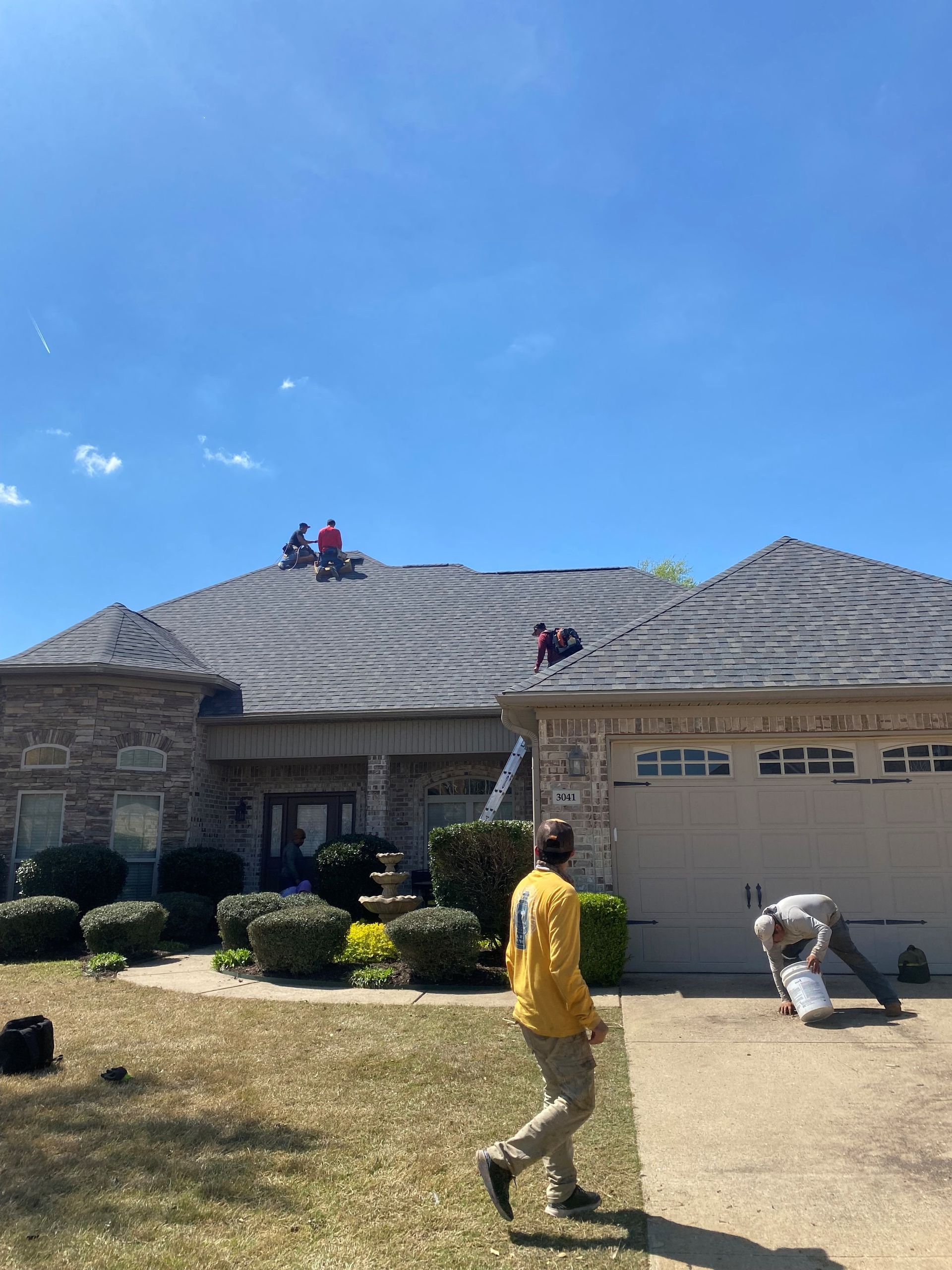 Workers on a roof, replacing shingles. One worker on the ground. Clear blue sky.
