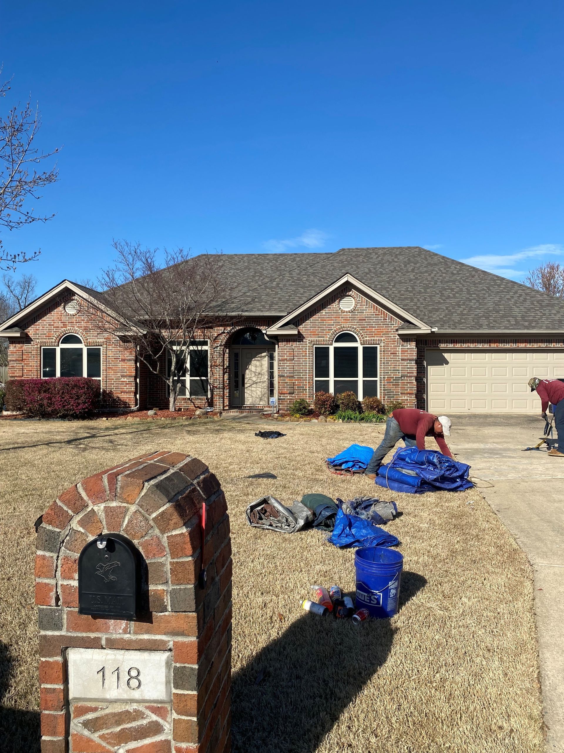 Brick house with people working on the lawn, blue tarps spread out. Blue sky. Mailbox.
