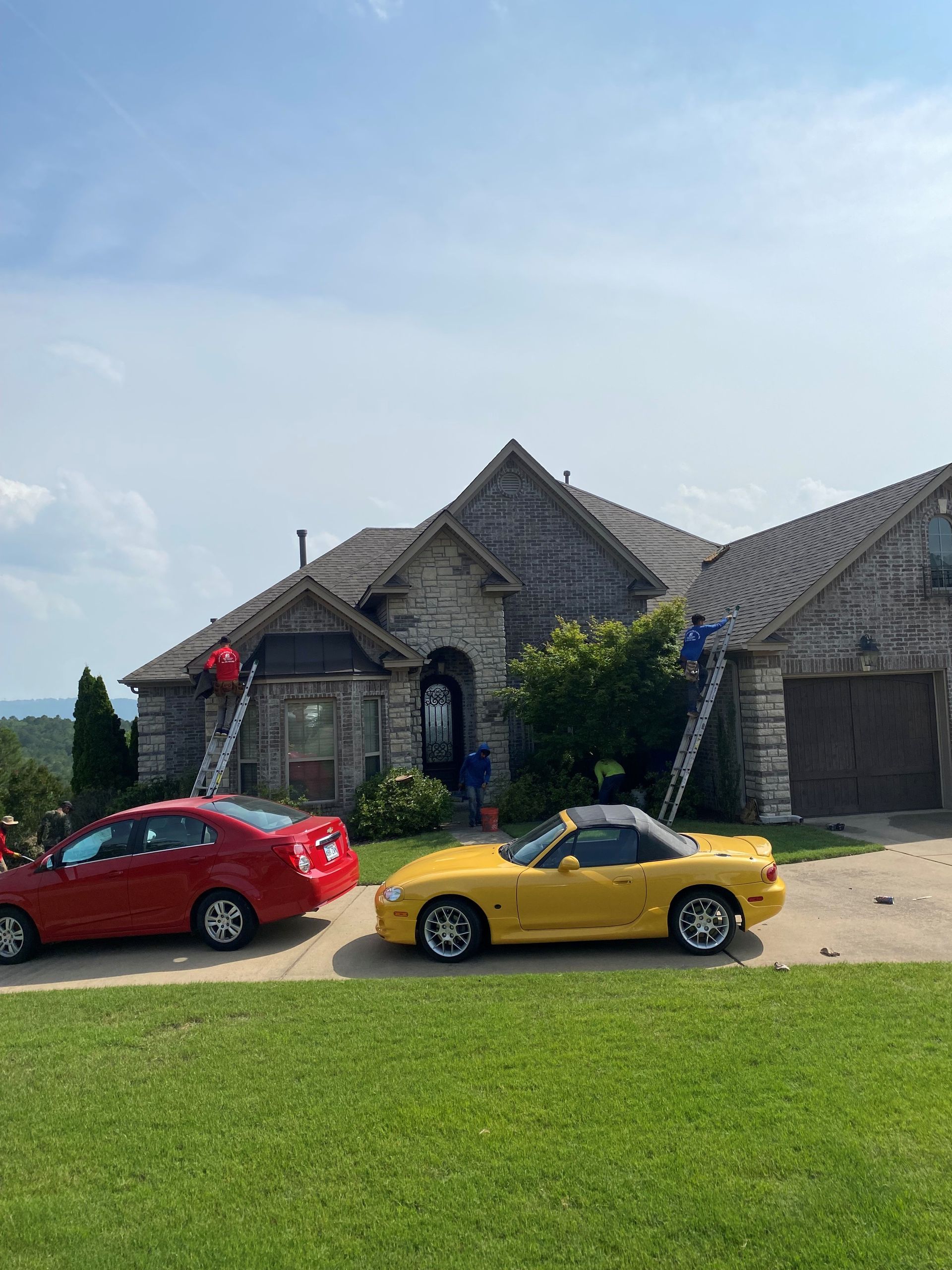 Two cars parked in front of a stone house with workers on ladders; blue sky. A red car is to the left, yellow convertible to the right.