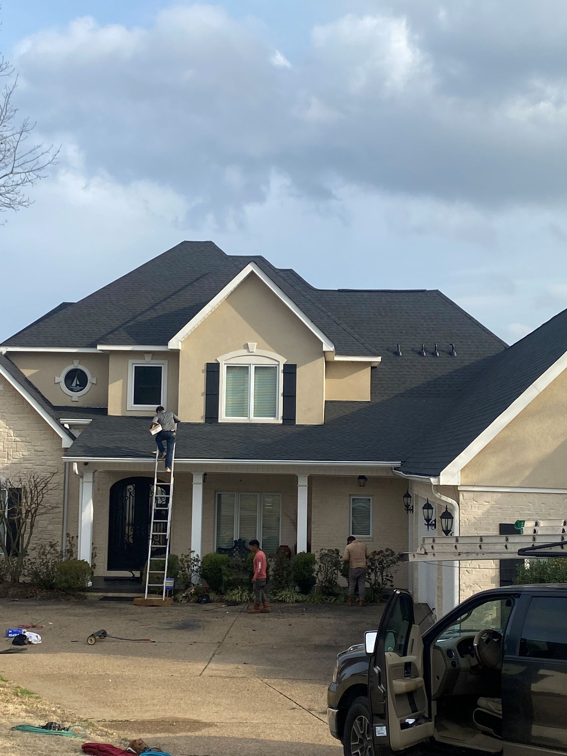 Workers on a roof with a black shingle roof, beige house, and a truck parked out front.
