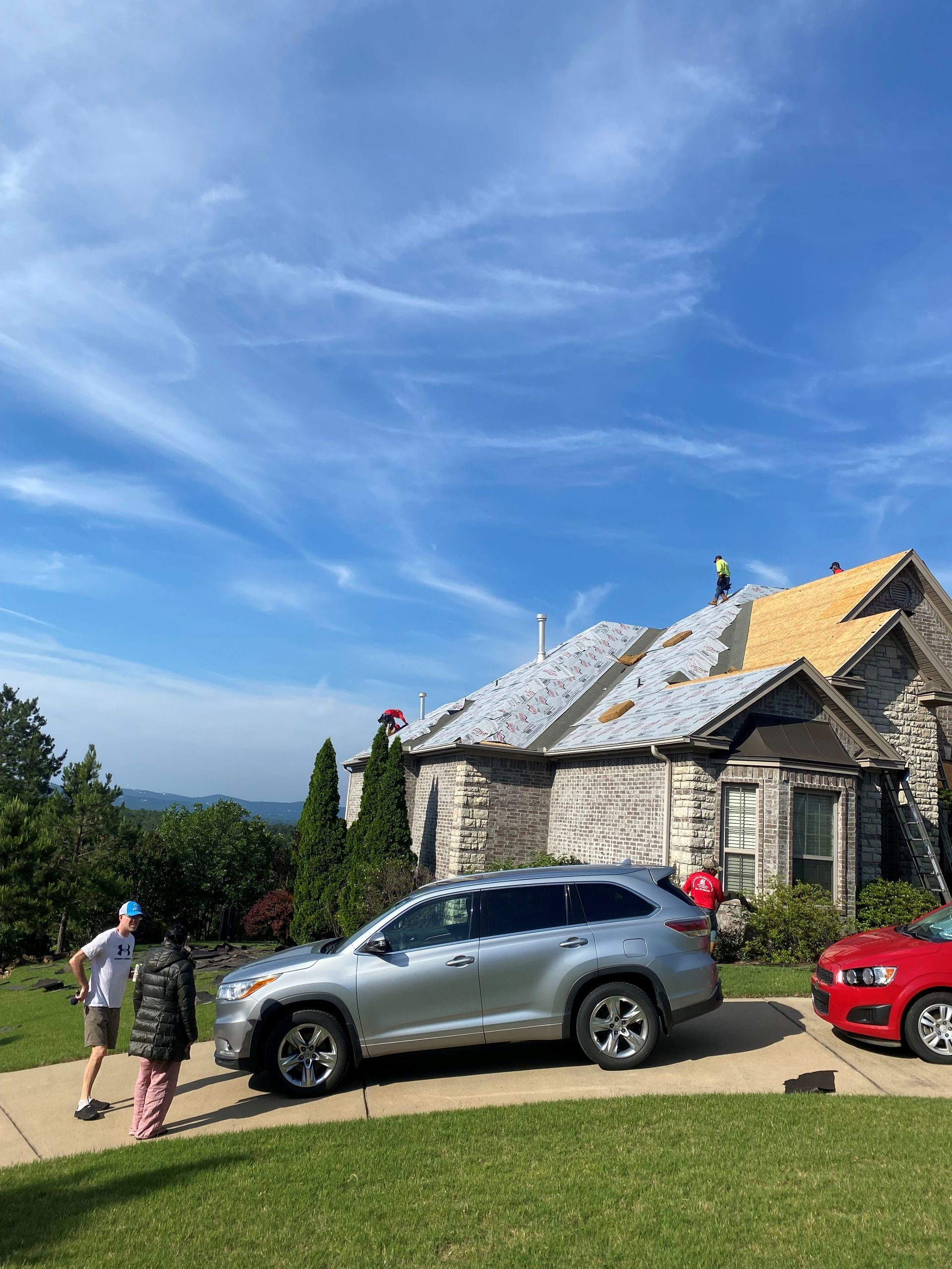 Silver SUV parked in front of a house with roofers working on the roof. People stand near the car on a sunny day.