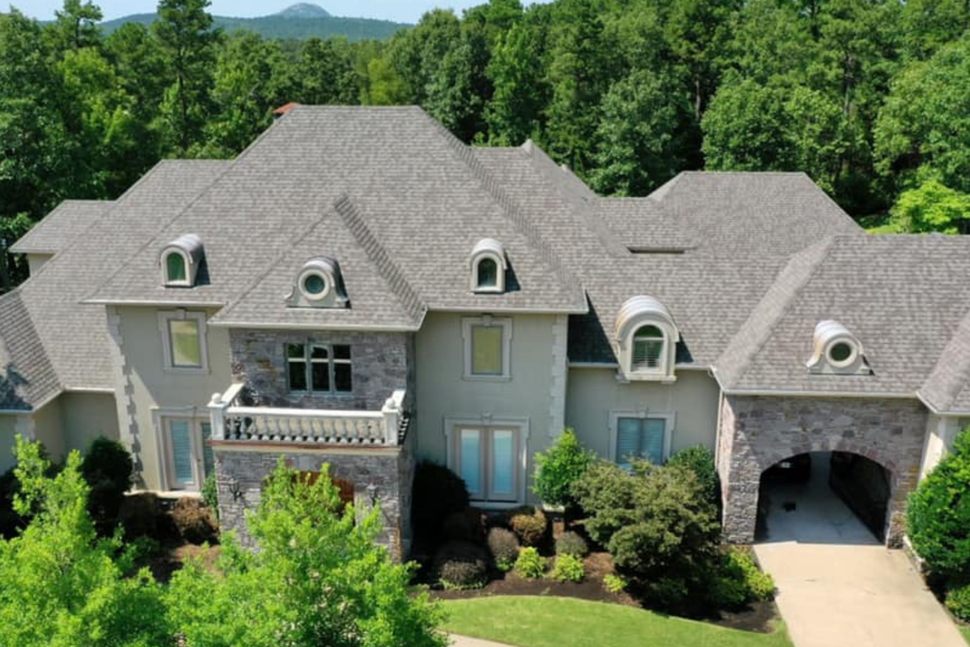 Large two-story house with gray roof, stone accents, and arched entry, set among lush green trees.