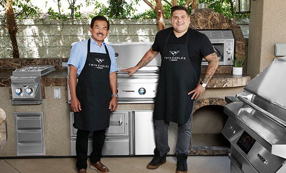 Two men wearing aprons are standing in front of a grill.