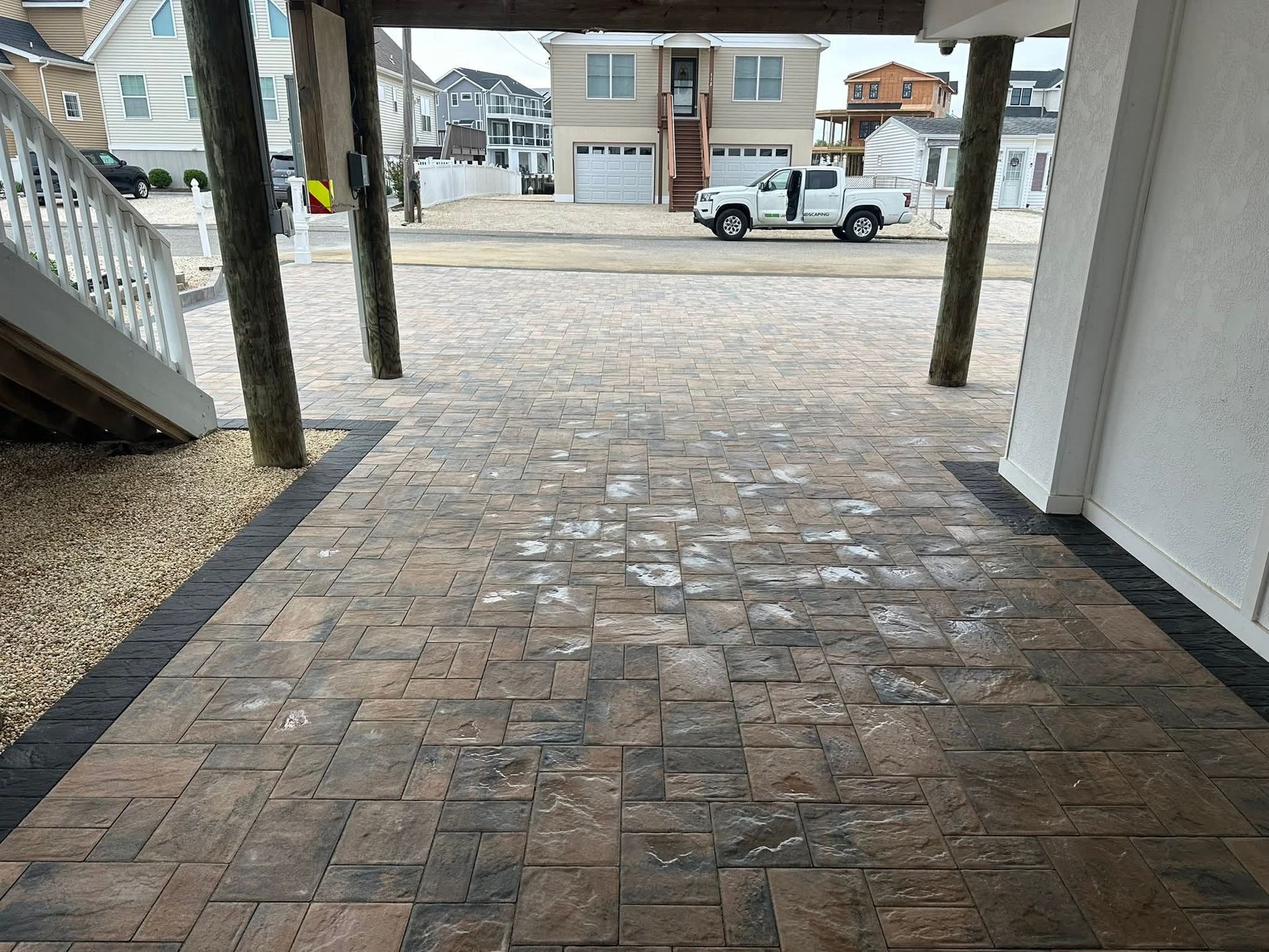Brick patio under a building, with a gravel border, leading to a street with houses and a truck.