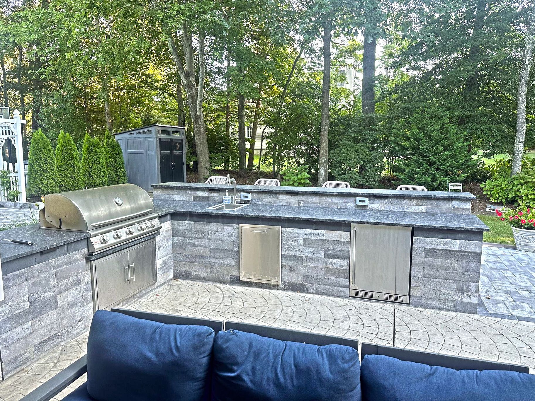 Outdoor kitchen with grill, storage, and seating on a patio, surrounded by greenery.