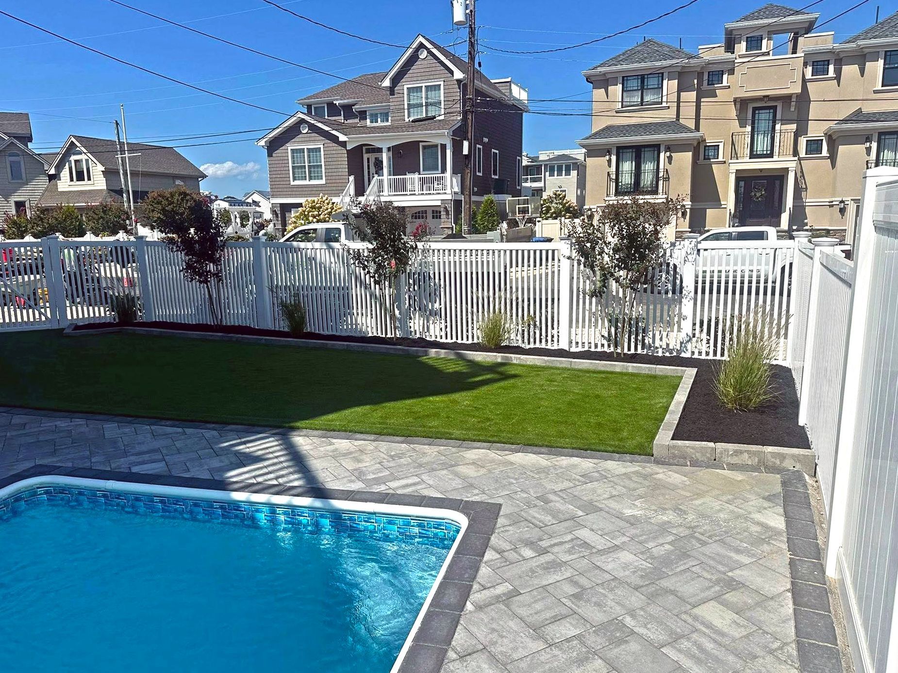 Backyard with a swimming pool, lawn, white picket fence, and houses in the background.