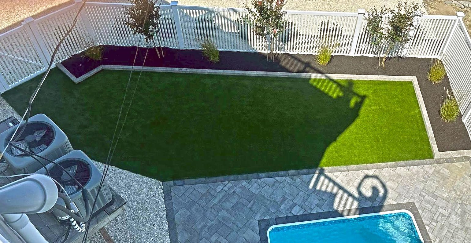 Overhead view of a backyard with green turf, black mulch beds, white fence, and pool.