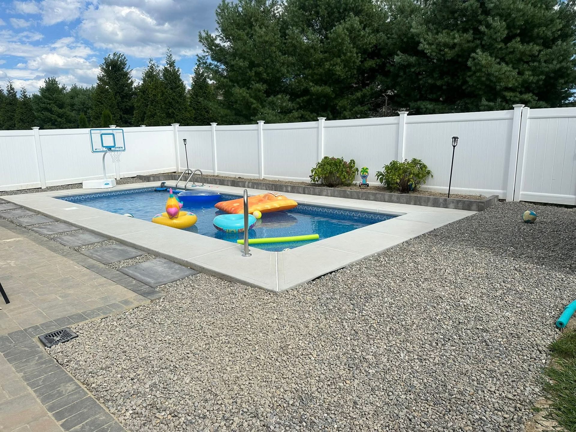 Rectangular pool with floats surrounded by gravel, concrete, and a white fence. Trees and sky visible.