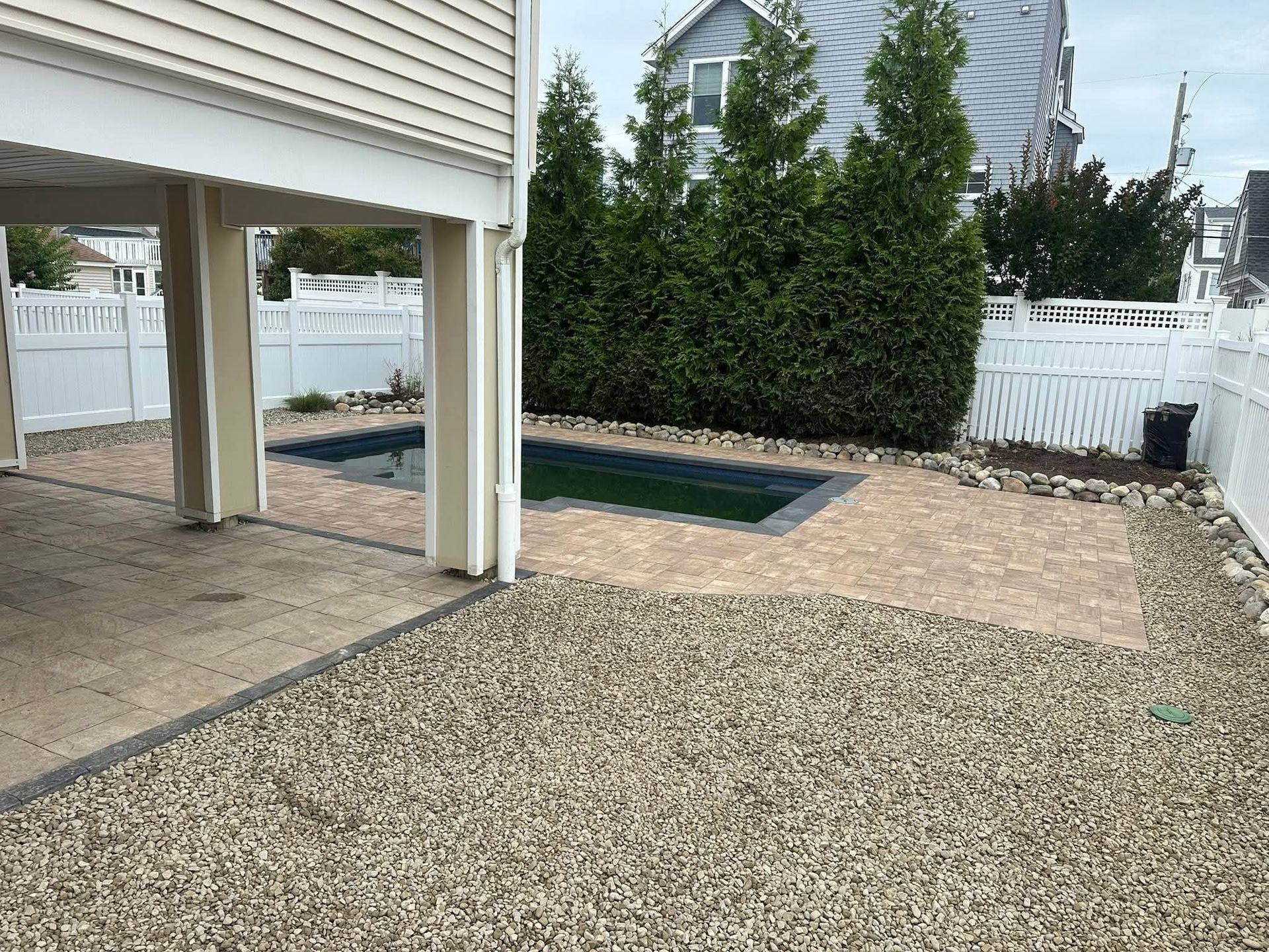 Backyard with a pool, gravel, and a white fence, under a raised house.