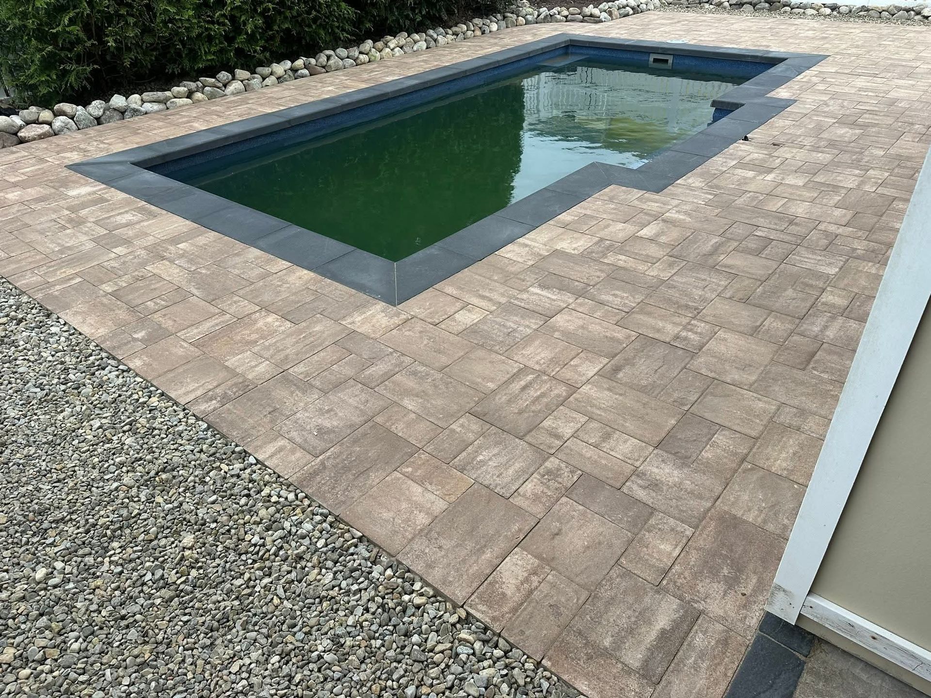 A rectangular pool with green water, surrounded by stone pavers, gravel, and a beige wall.
