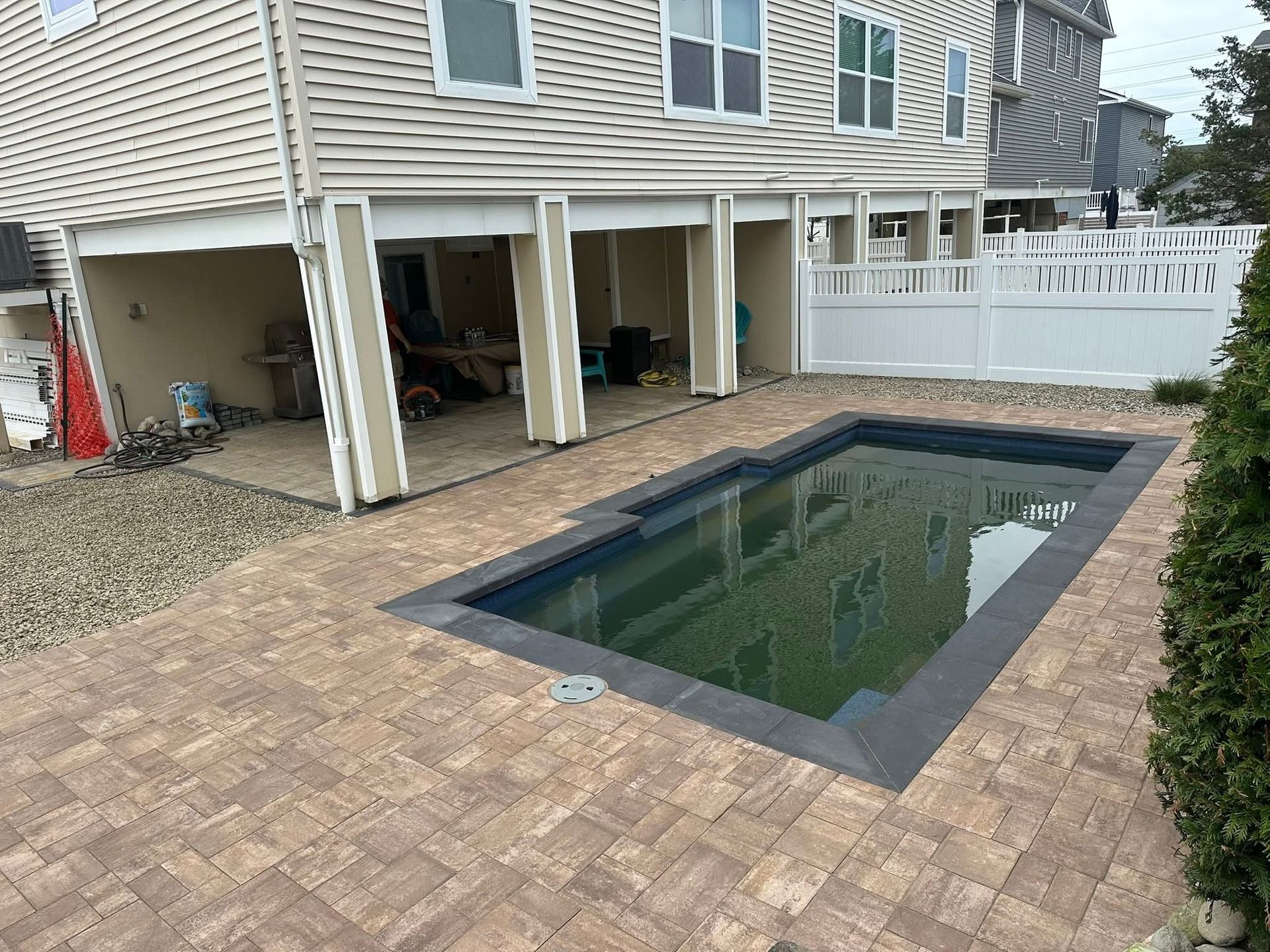 A rectangular pool with dark gray trim, surrounded by tan pavers, under a beige elevated house.