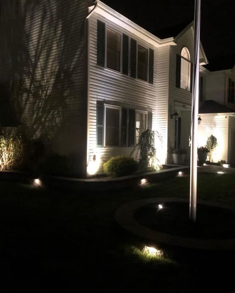 Two-story house at night illuminated by landscape lighting. White siding, black shutters, and a flag pole are visible.
