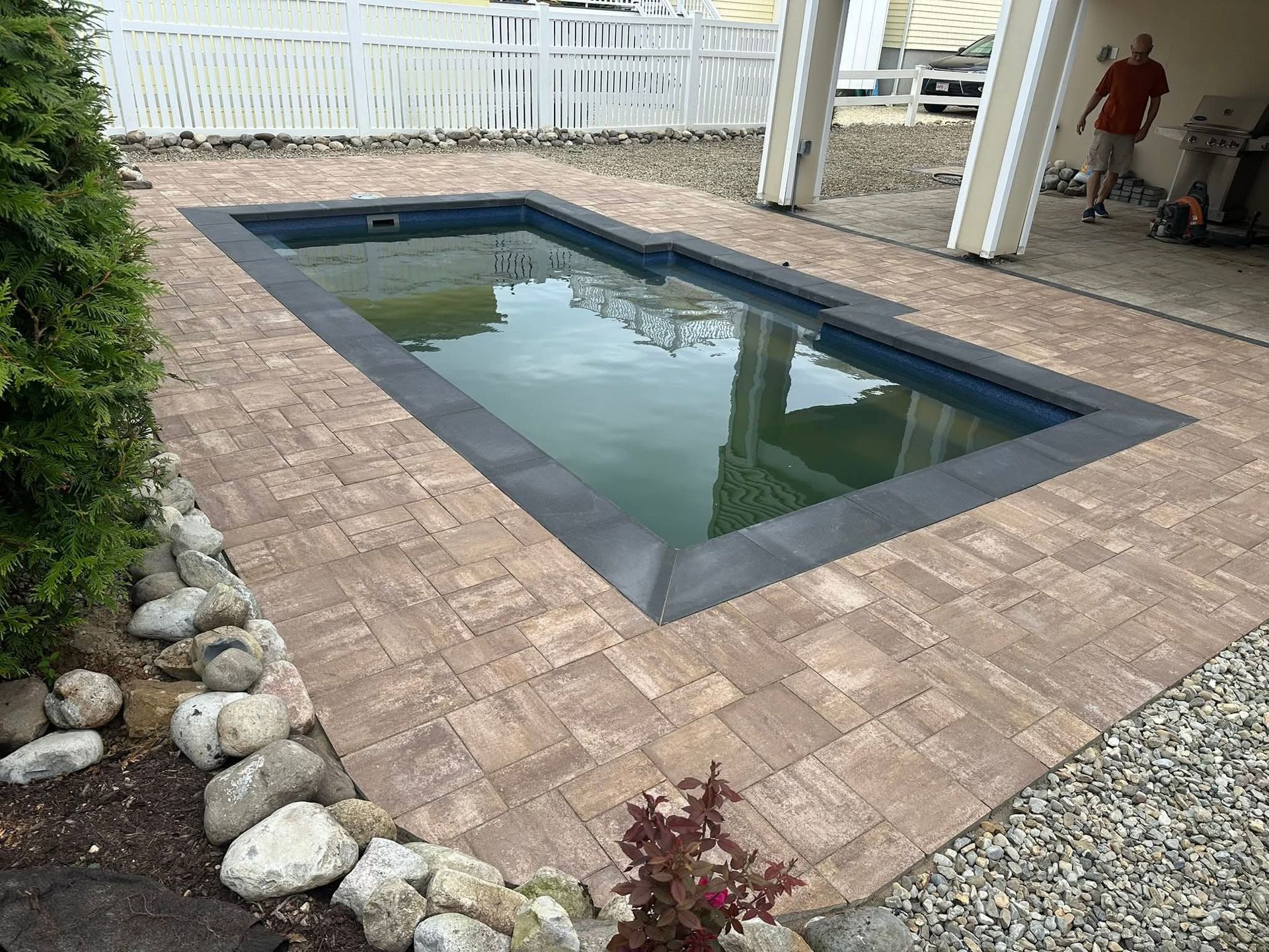 Small rectangular pool with dark coping, surrounded by brown pavers, next to a yard with rocks and a white fence.