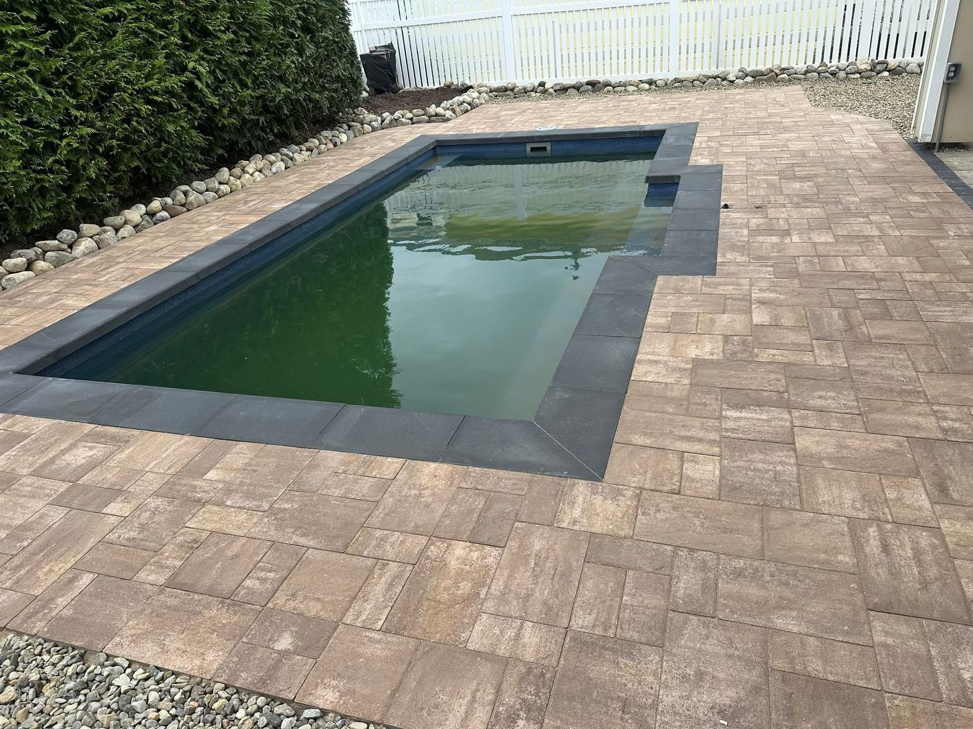 Rectangular outdoor pool surrounded by brown pavers and a white fence; water is green.