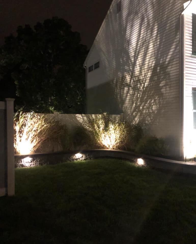 Lawn lit by ground lights; building on the right, tree casting shadows on wall.