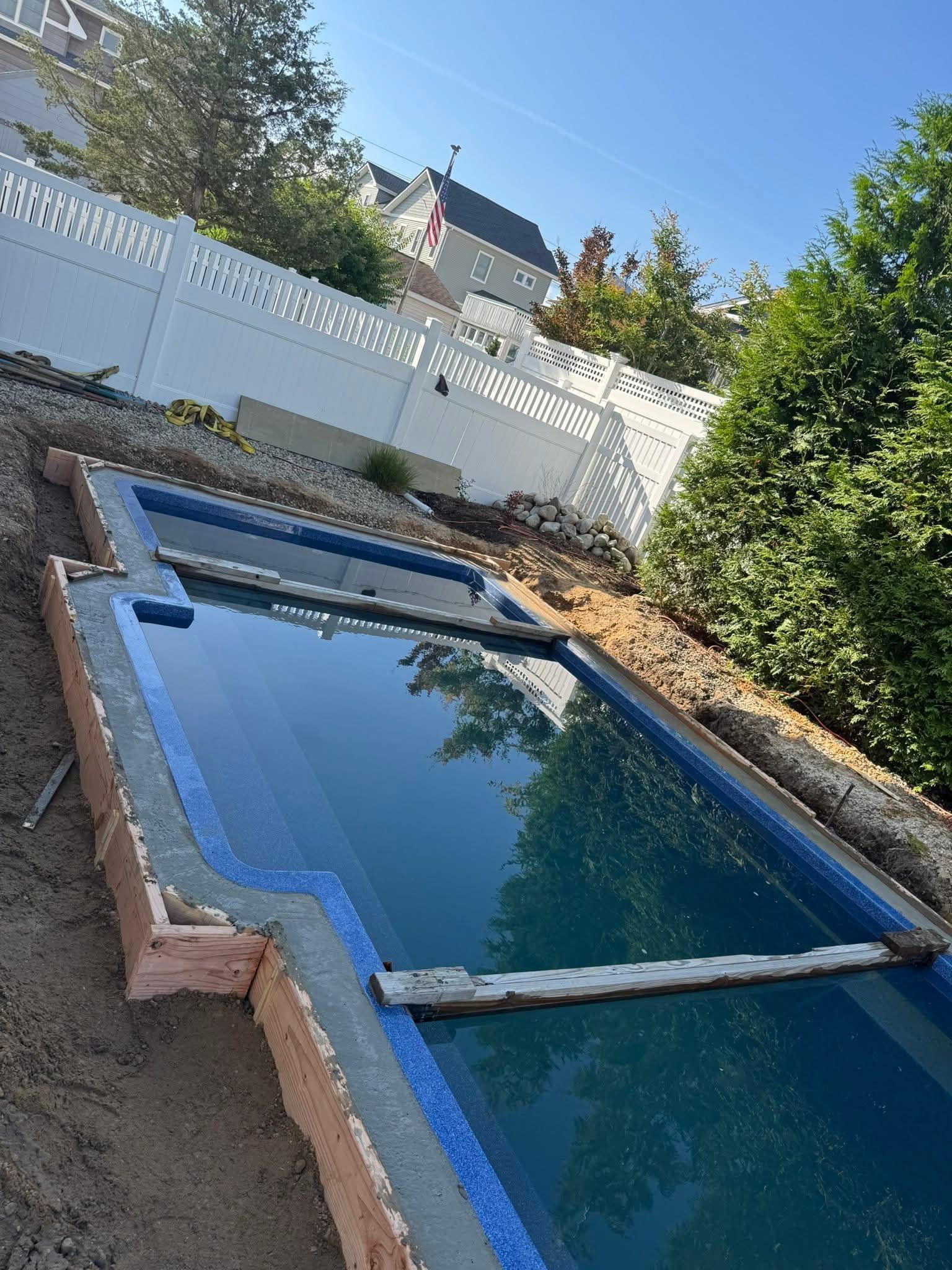 Pool under construction with blue lining, wood planks, and surrounding soil. White fence and trees in the background.