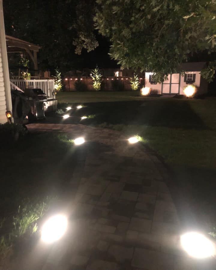 Brick pathway illuminated by lights at night, leading to a lit shed in a yard.