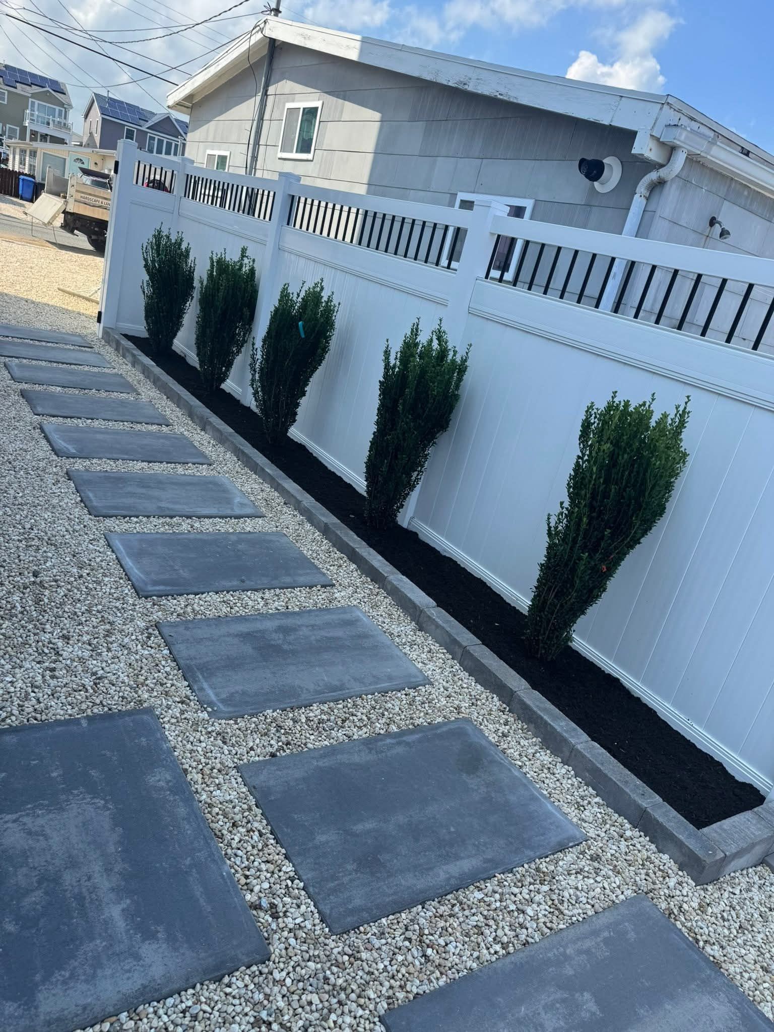 A paved pathway with gray square stones and gravel, leading towards a white fence with trimmed green bushes.