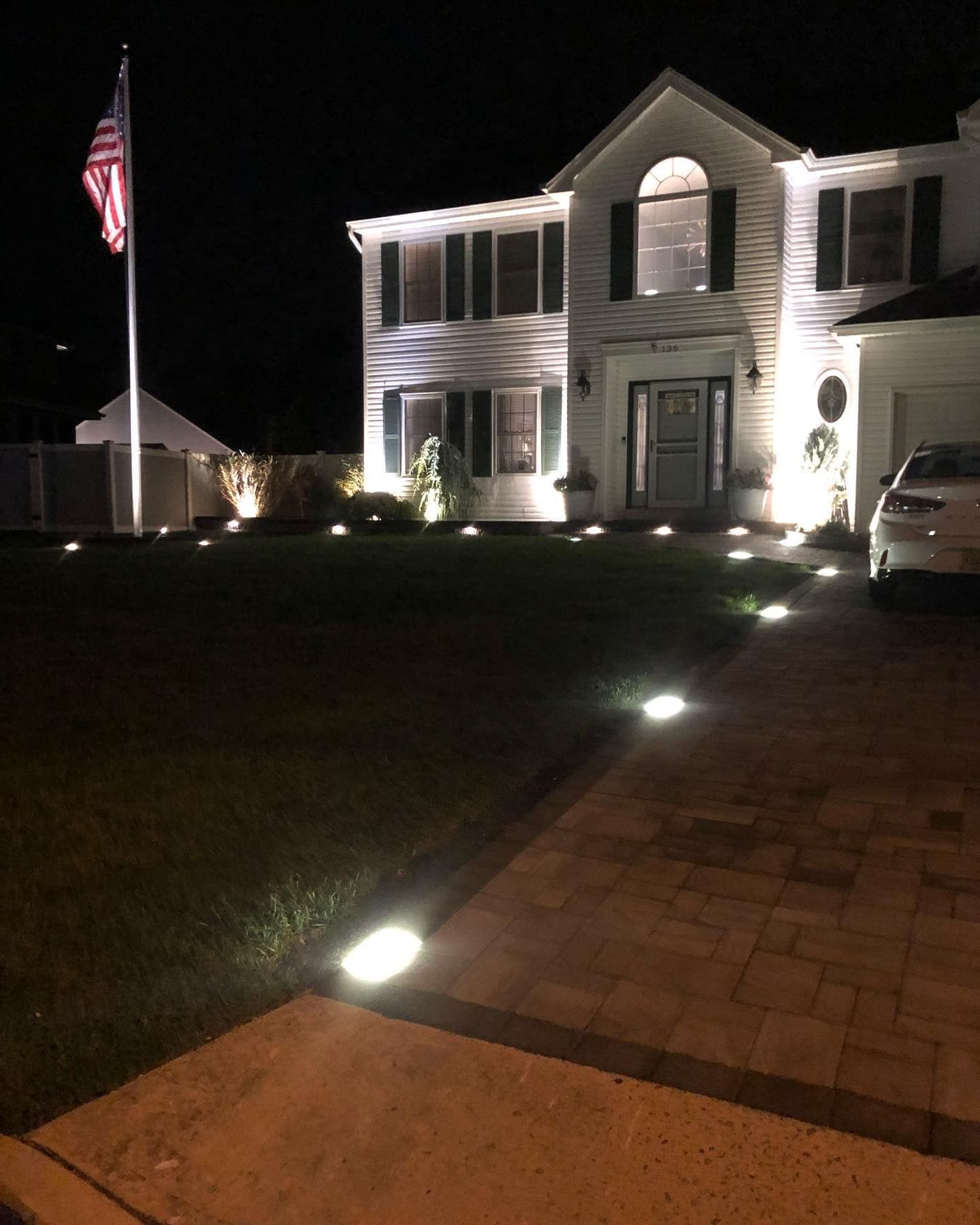 Two-story white house at night, lit by spotlights on lawn and driveway. American flag flies.
