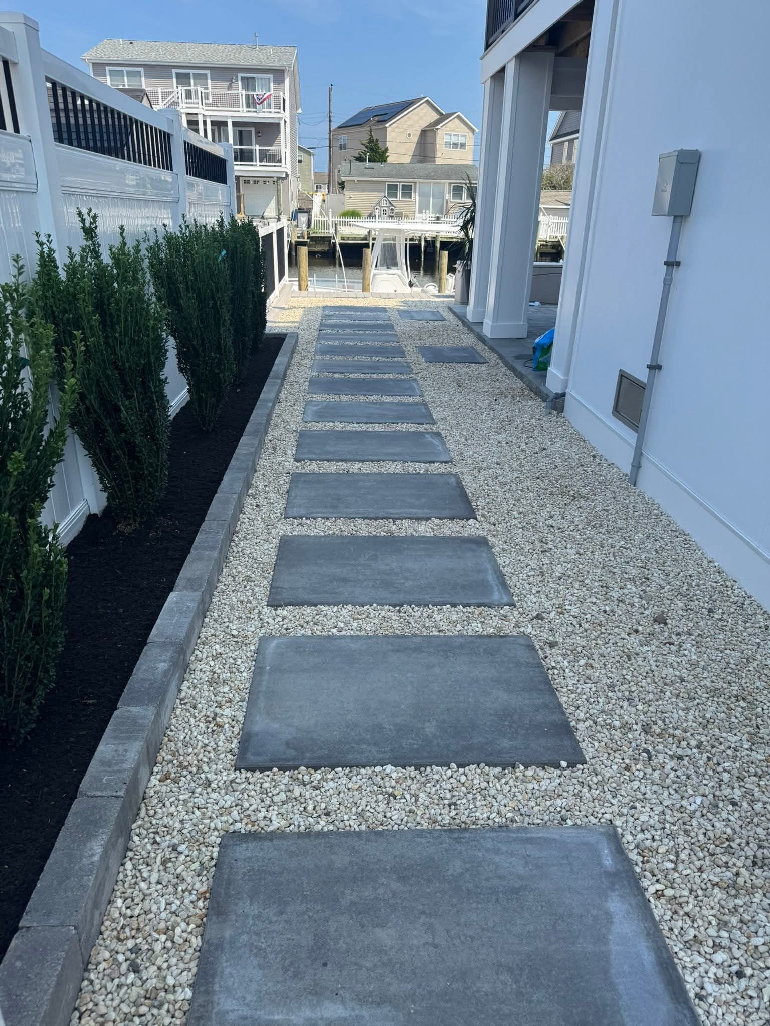 Stone pavers create a walkway between a building and hedges filled with white gravel.