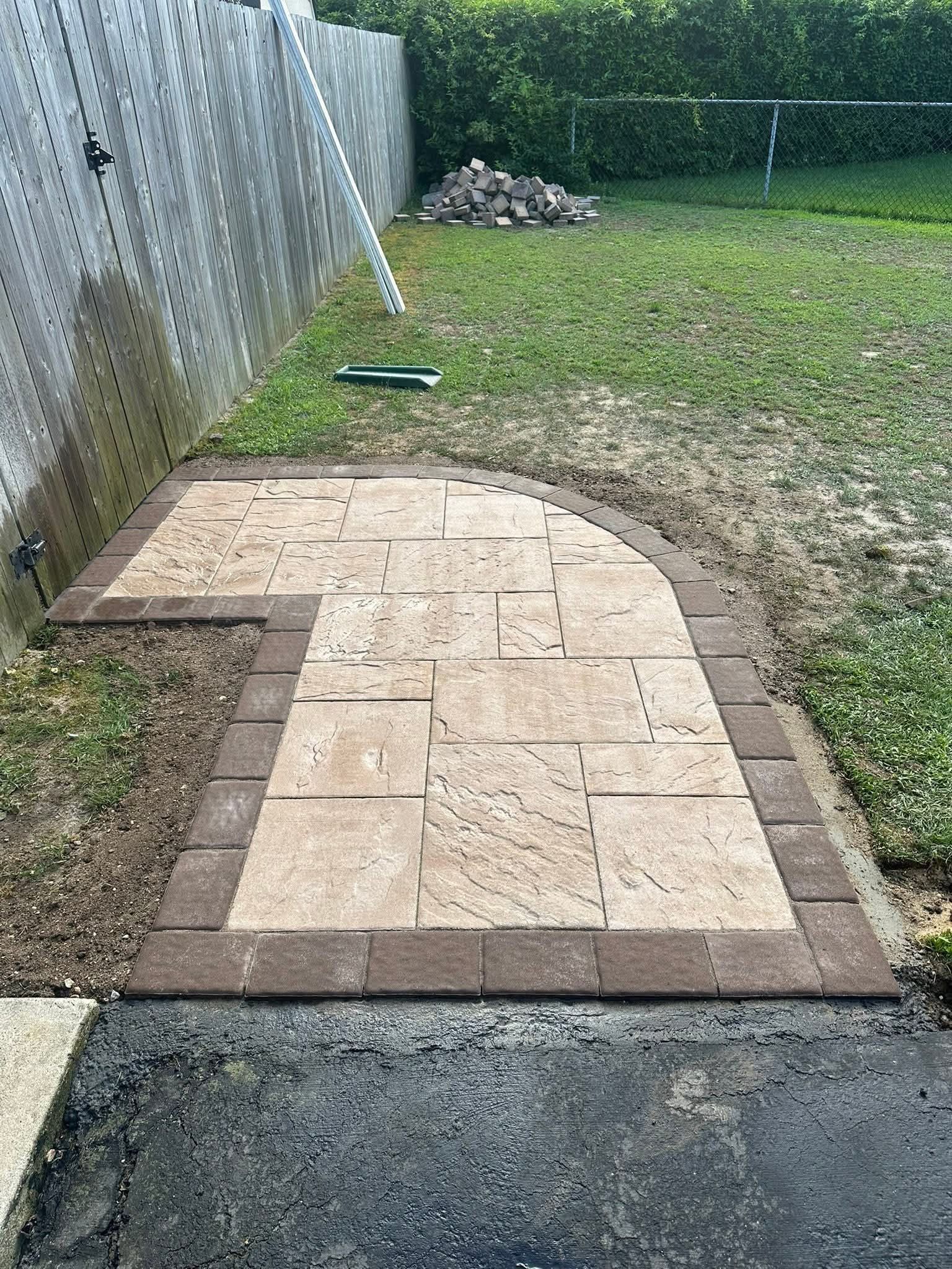 Paved patio with brown edging, next to a wooden fence and grassy yard.