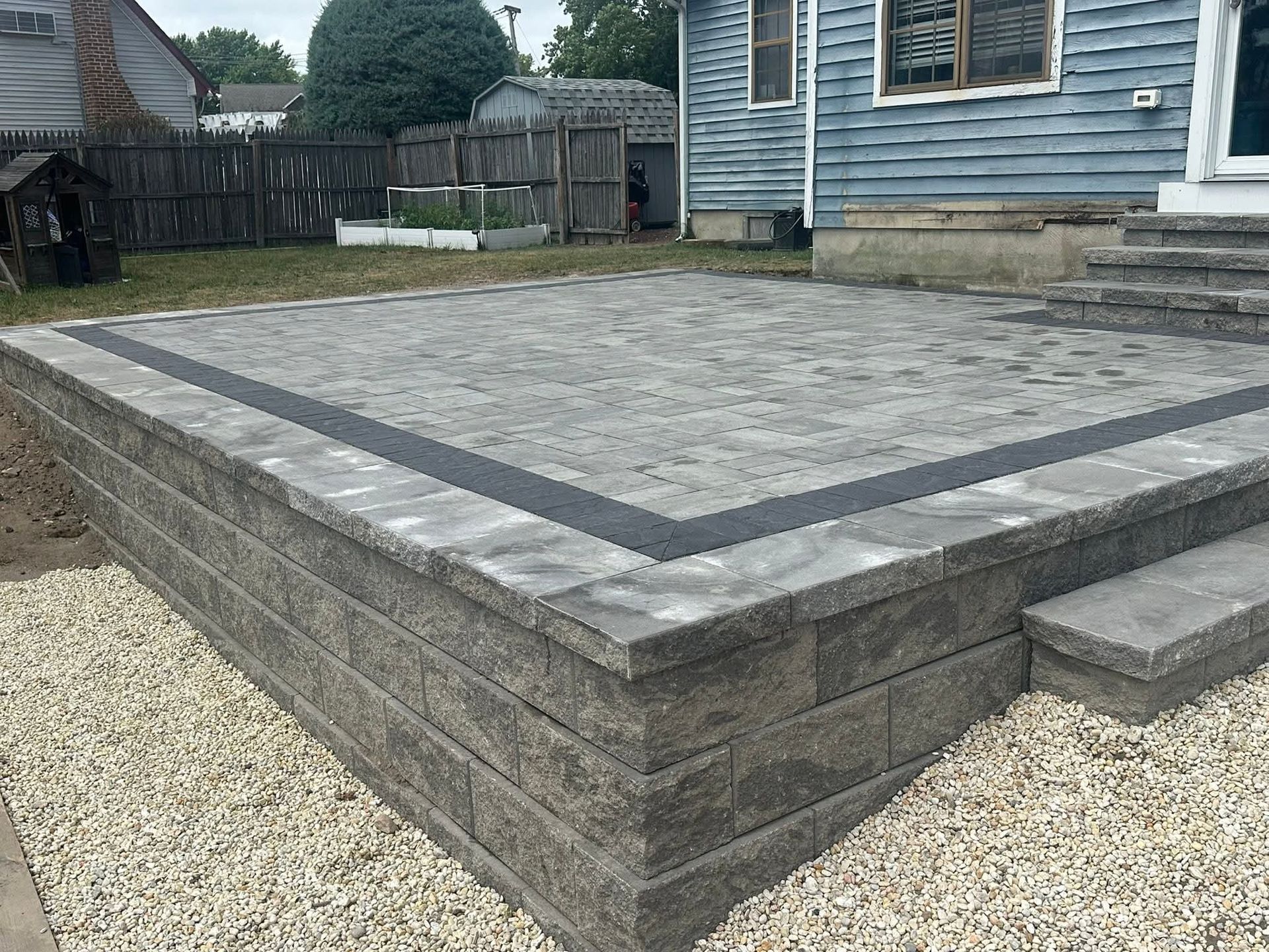 Gray brick patio with retaining wall, pea gravel, and blue house.