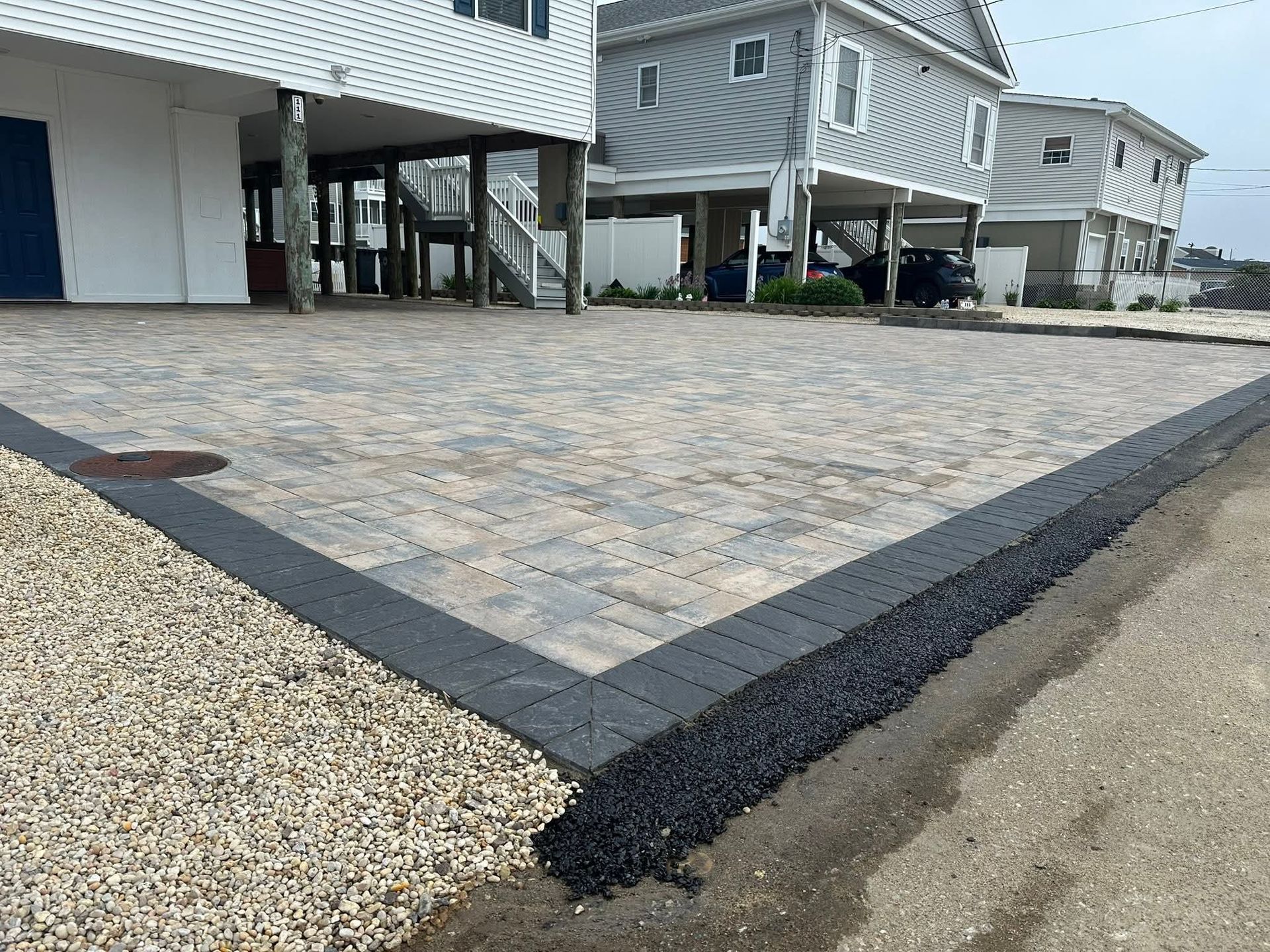 Paver driveway bordered by dark pavers, with gravel edge next to street and elevated houses.