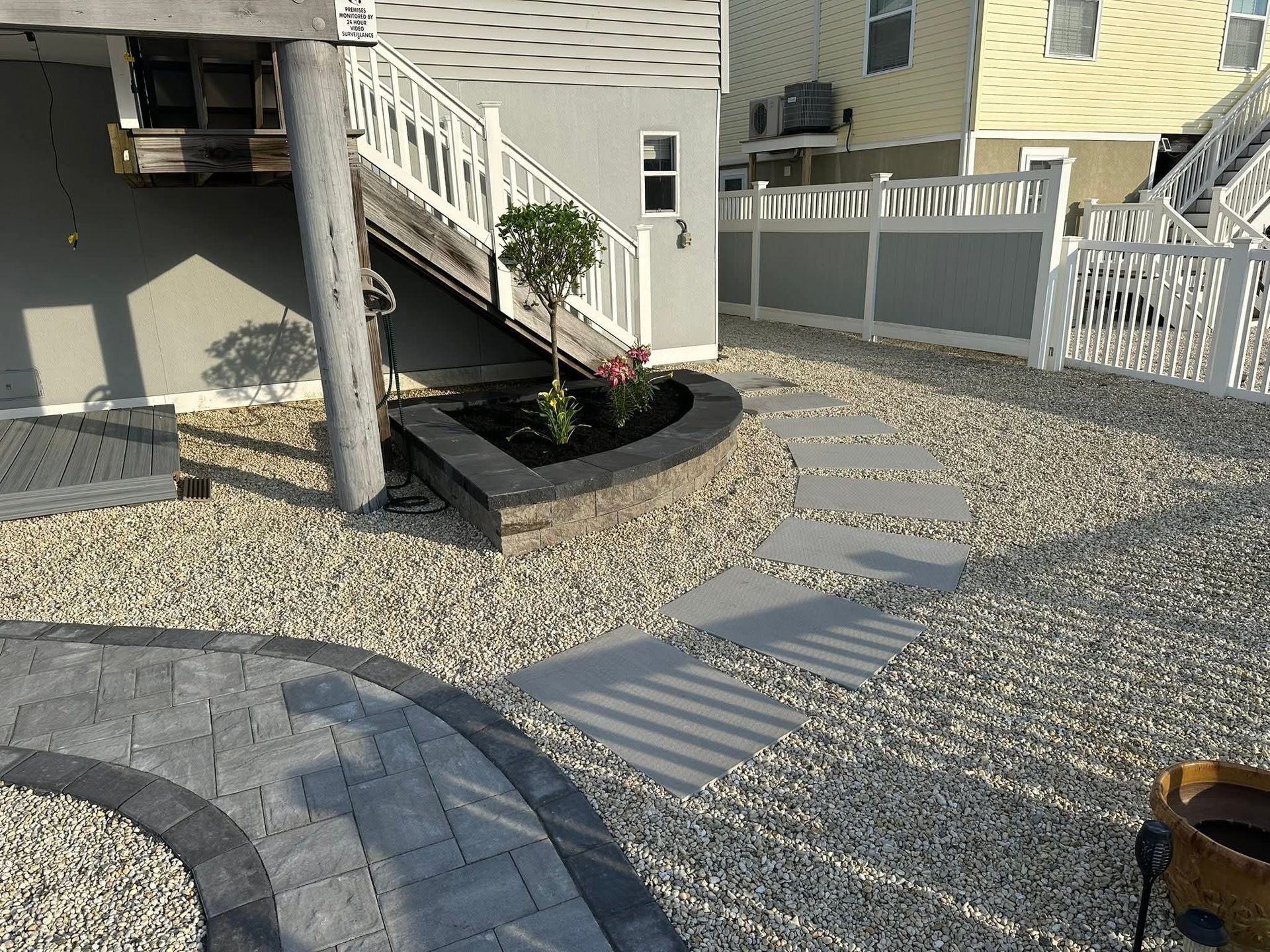 A gravel yard with stepping stones, a curved paved walkway, a small flower bed, and a white fence.