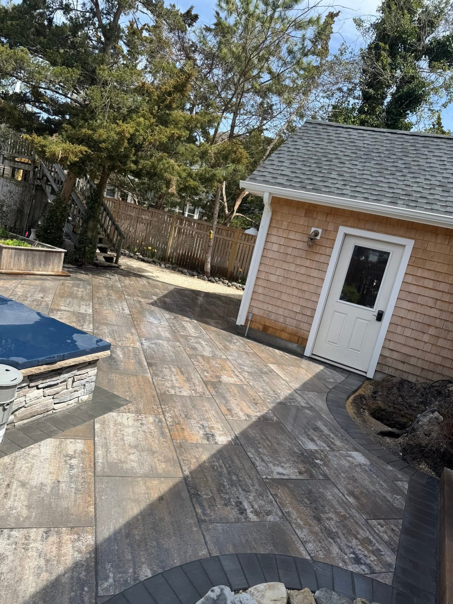 Patio with multi-toned paving stones, small tan shed with white door. Trees in background, sunny day.