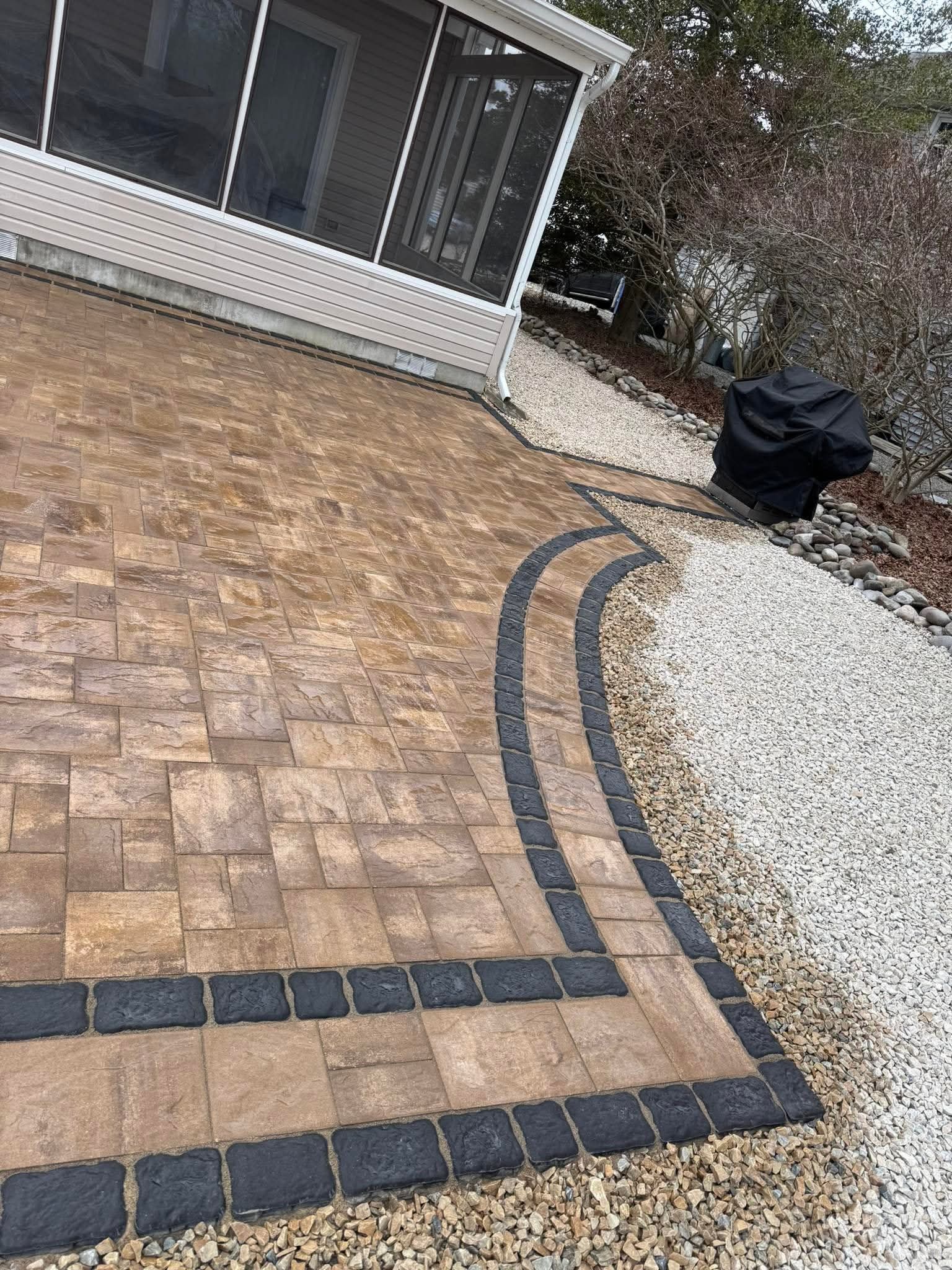 Patio with tan pavers and black border, gravel path, and covered grill.