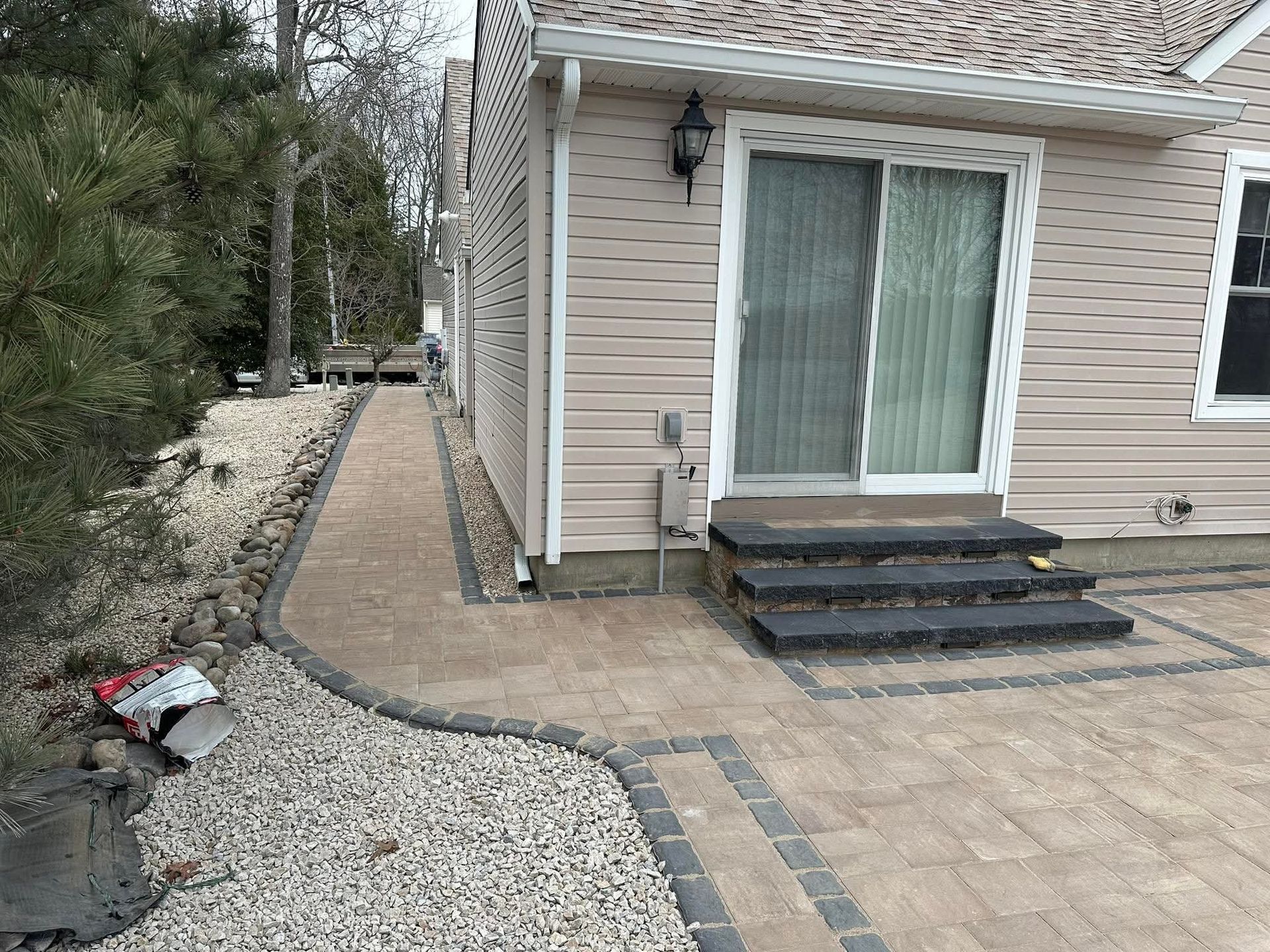 Paver pathway and patio outside a beige-sided house with steps leading to a sliding door; gravel border and landscaping.