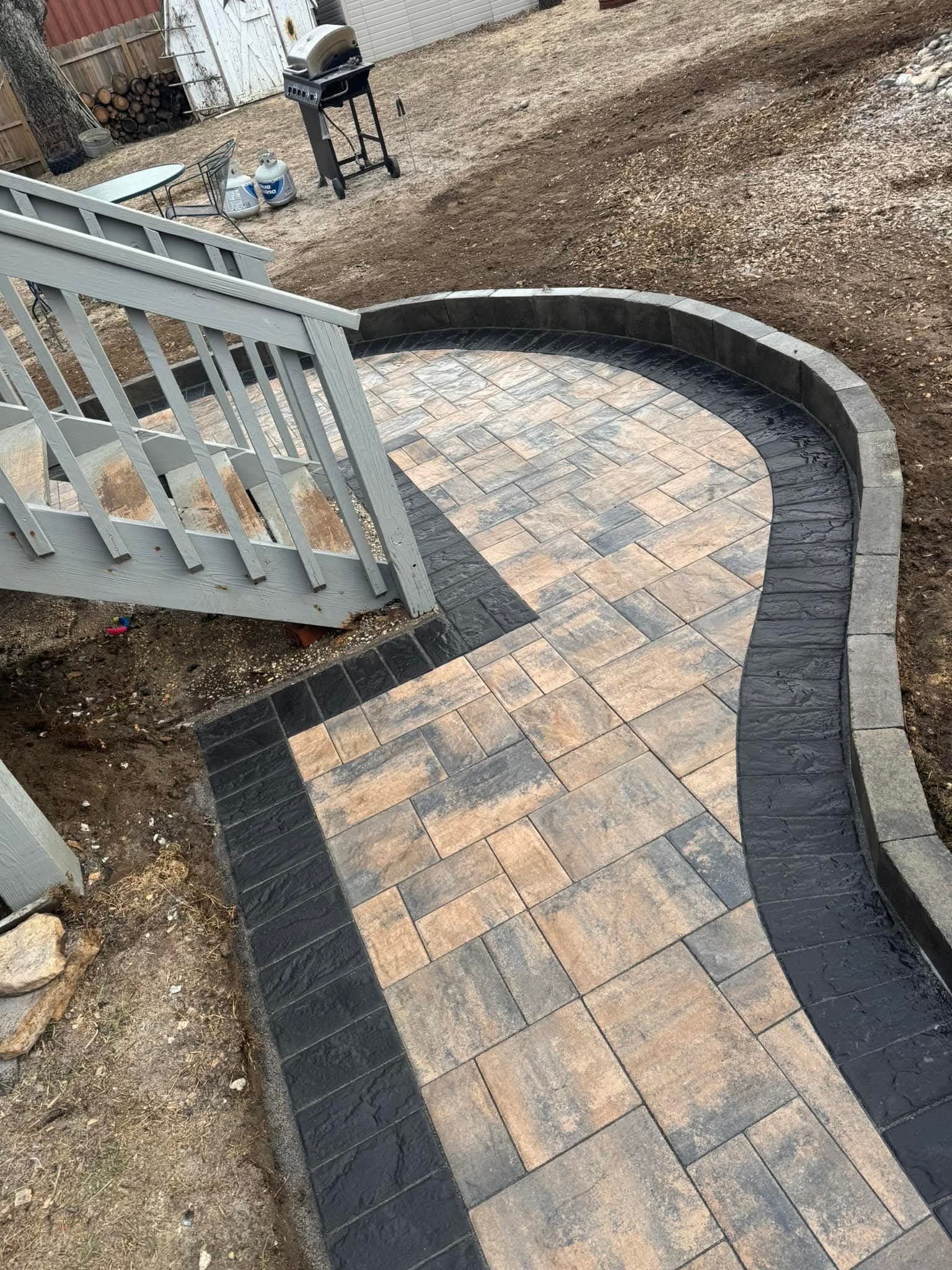 Brick pathway curves from stairs, bordered by dark pavers and a retaining wall; outdoors.