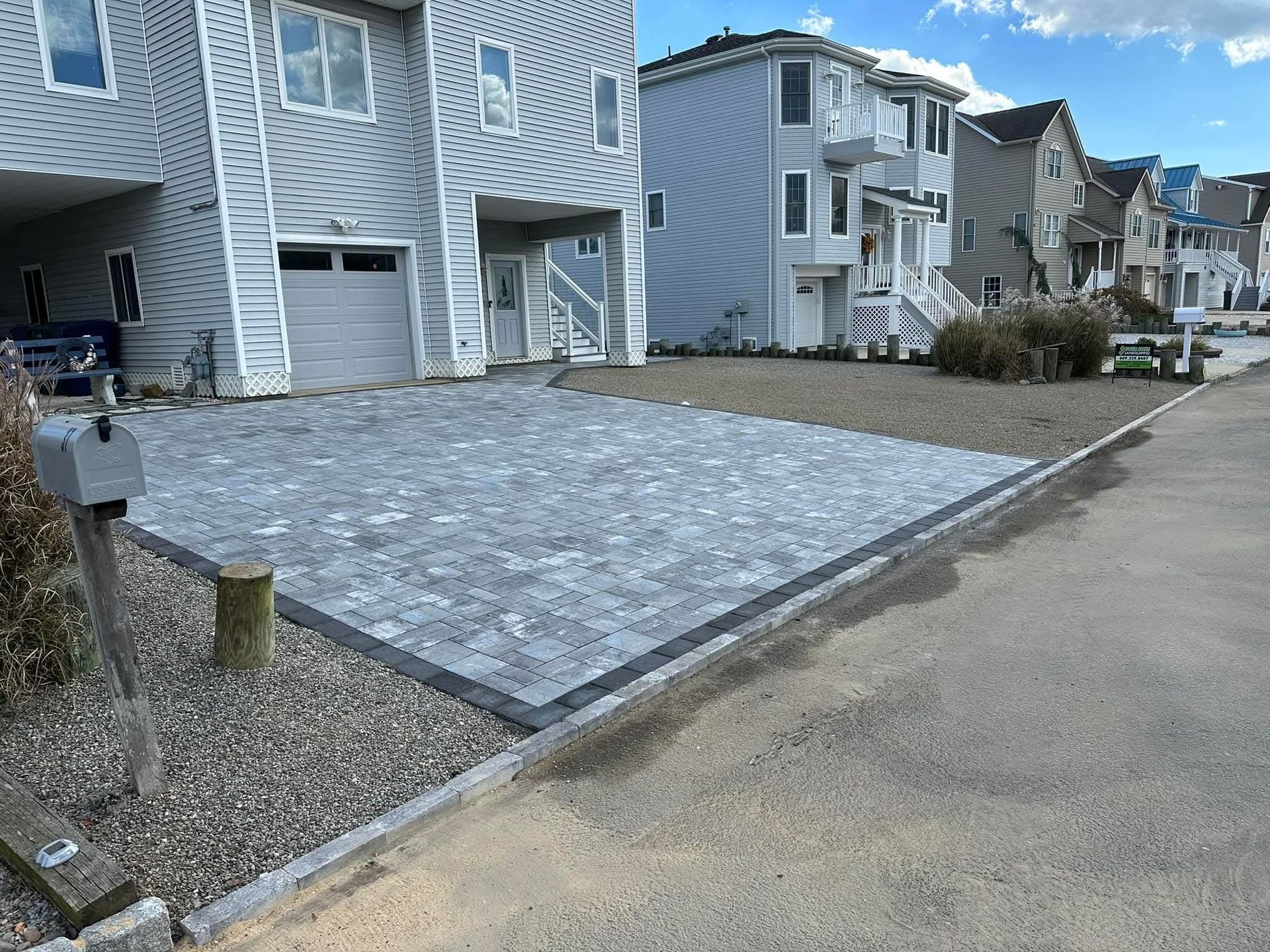 Driveway paved with gray blocks next to a street, bordered by a gray curb and gravel. Two-story houses in the background.