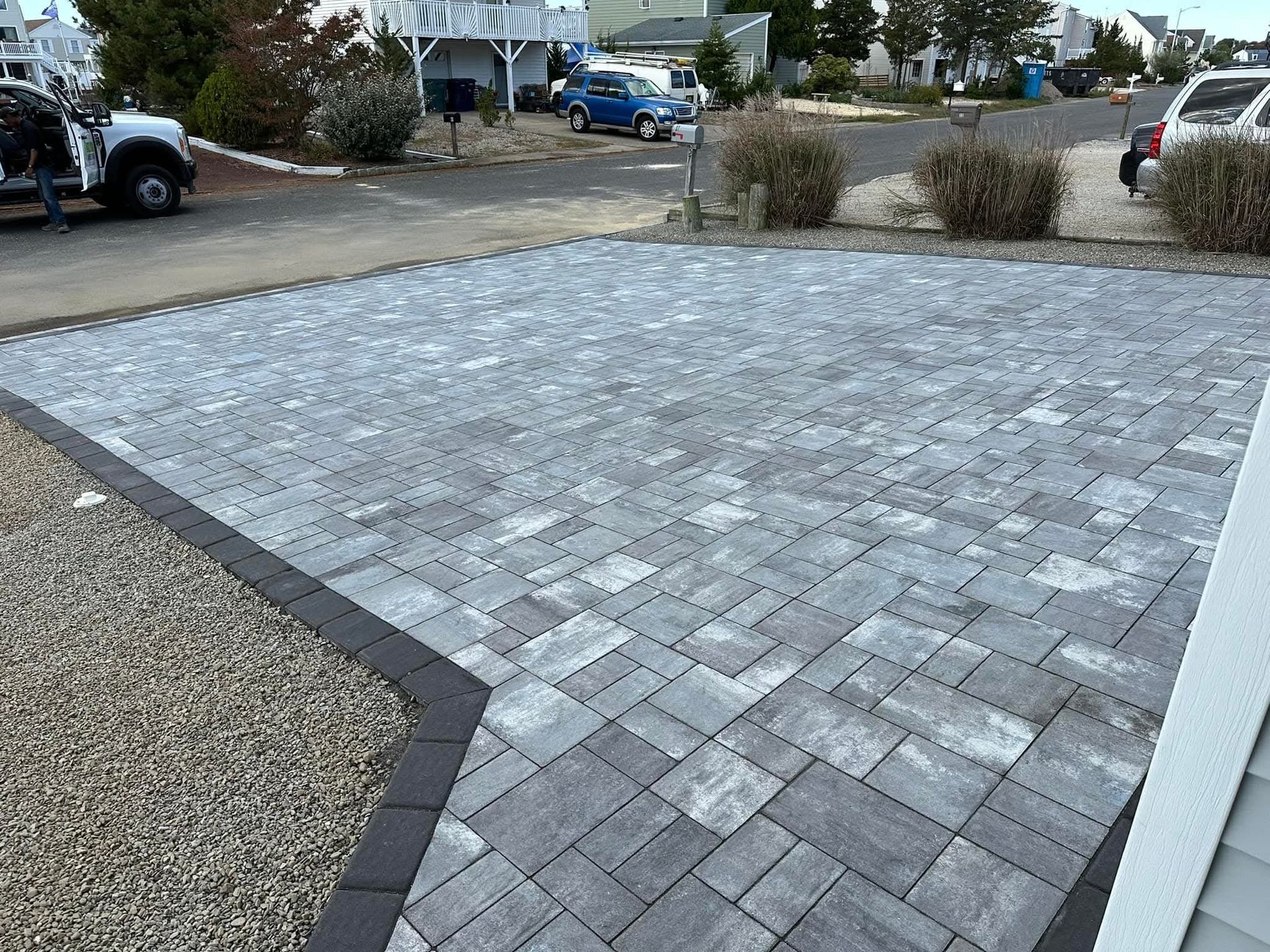 Brick pavers driveway in gray and black, next to gravel and a house. Cars in background.