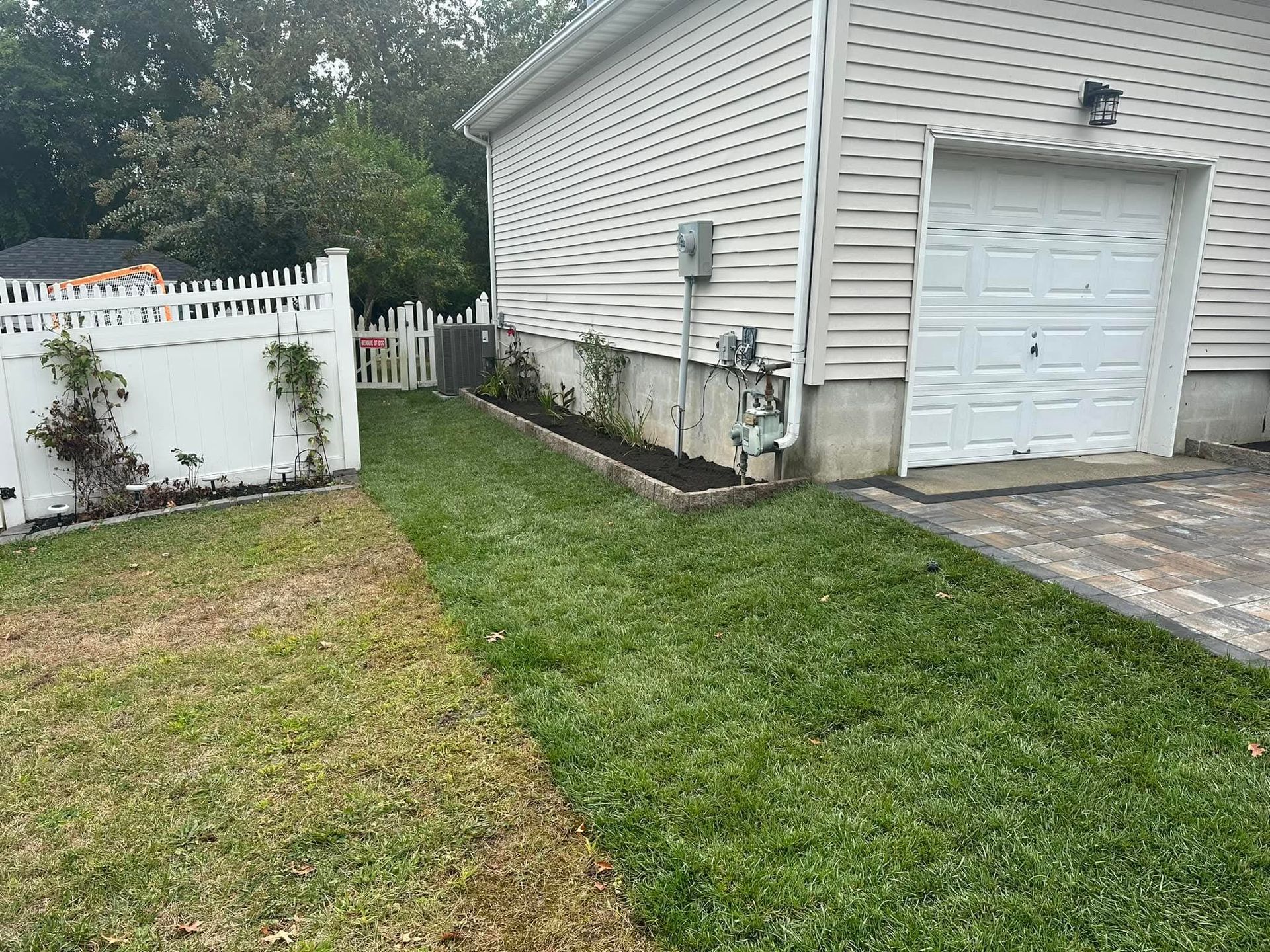 A side yard with green grass, a white fence, and a garage. New plantings are along the foundation of the house.