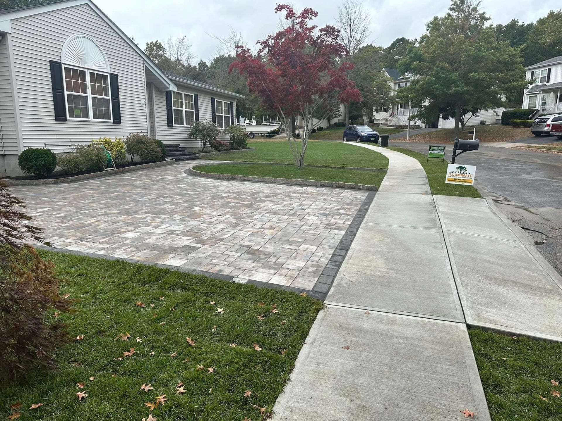 Brick driveway and walkway in front of a house with a small tree, grass, and parked cars.