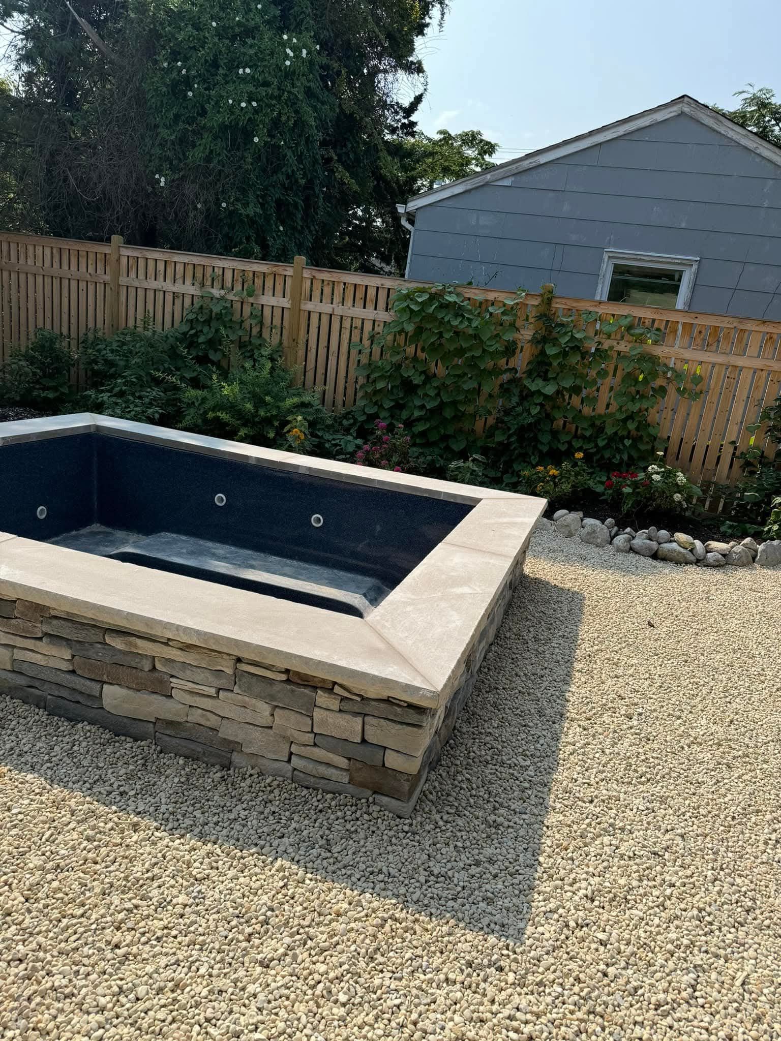 Stone-surrounded plunge pool in a backyard, surrounded by gravel, with a wooden fence and house in the background.