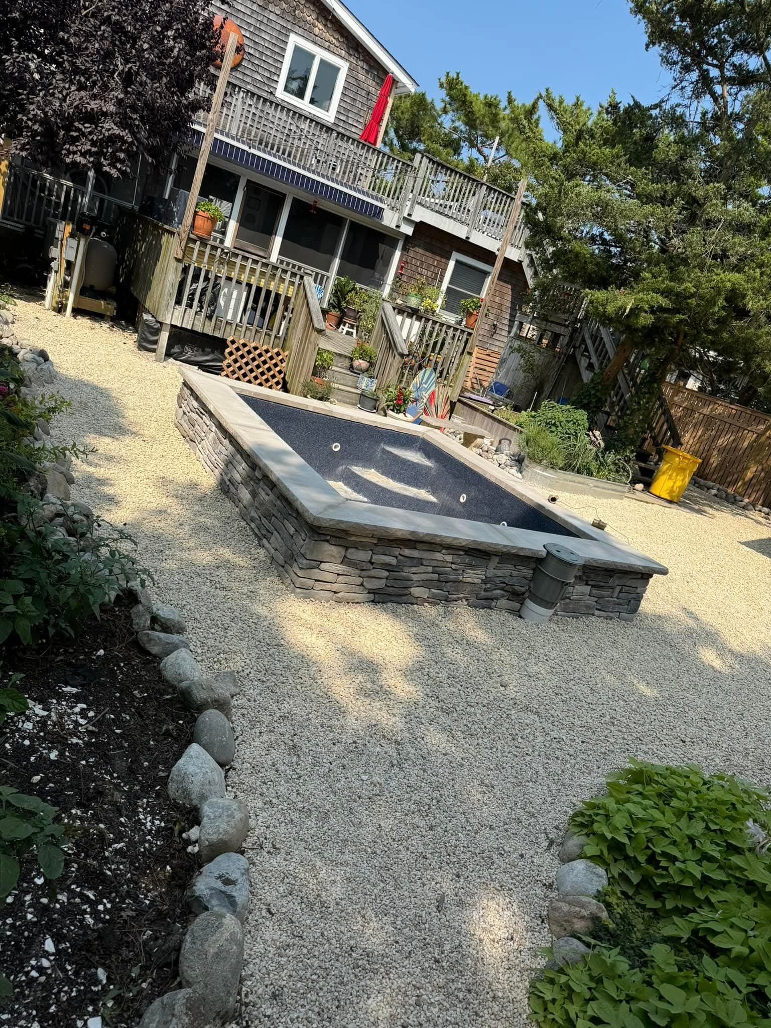 House with a stone fire pit on a gravel-covered slope, with landscaping and trees.