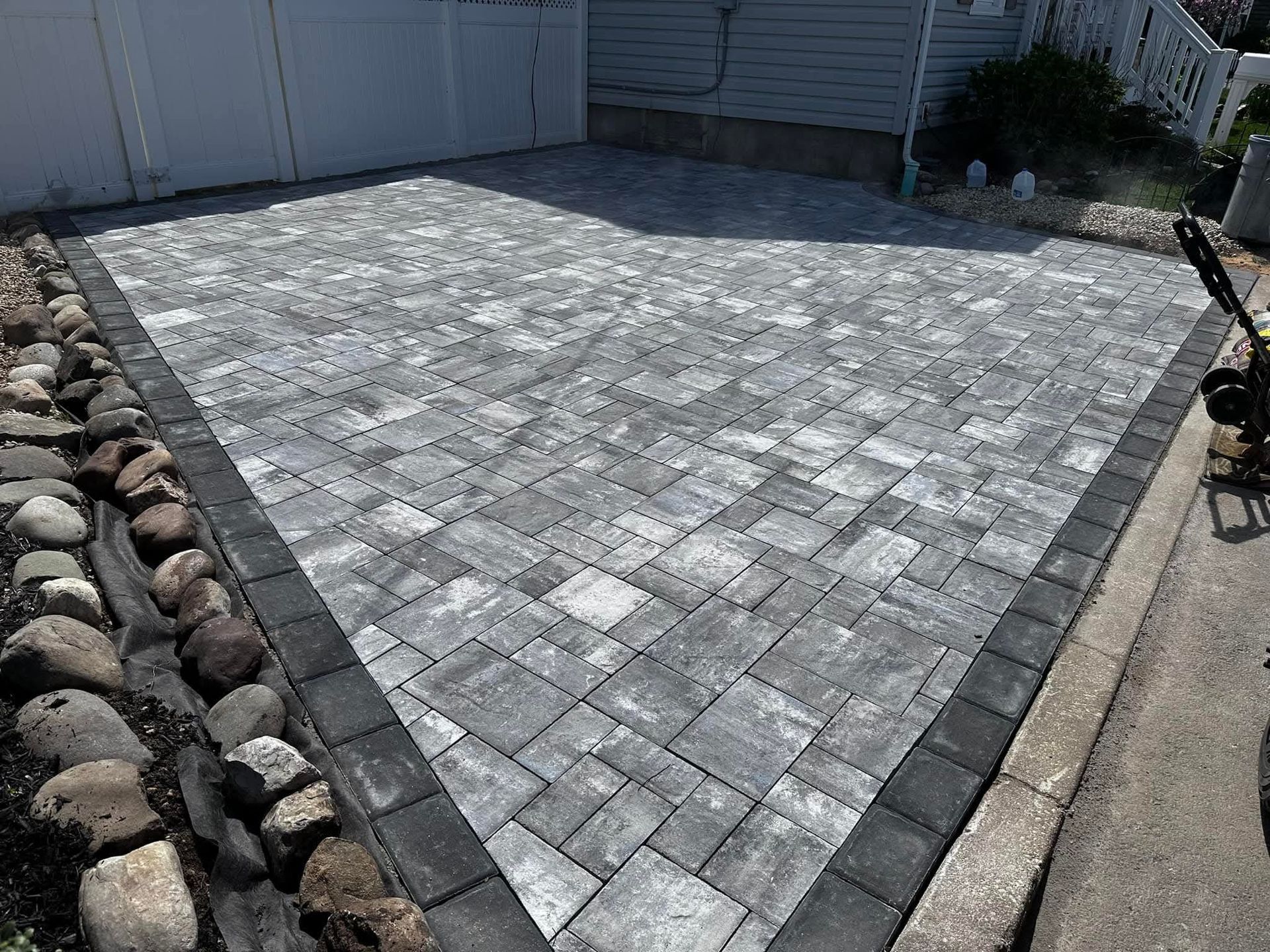 Gray brick driveway with black border and stone landscaping next to white fence and a concrete curb.