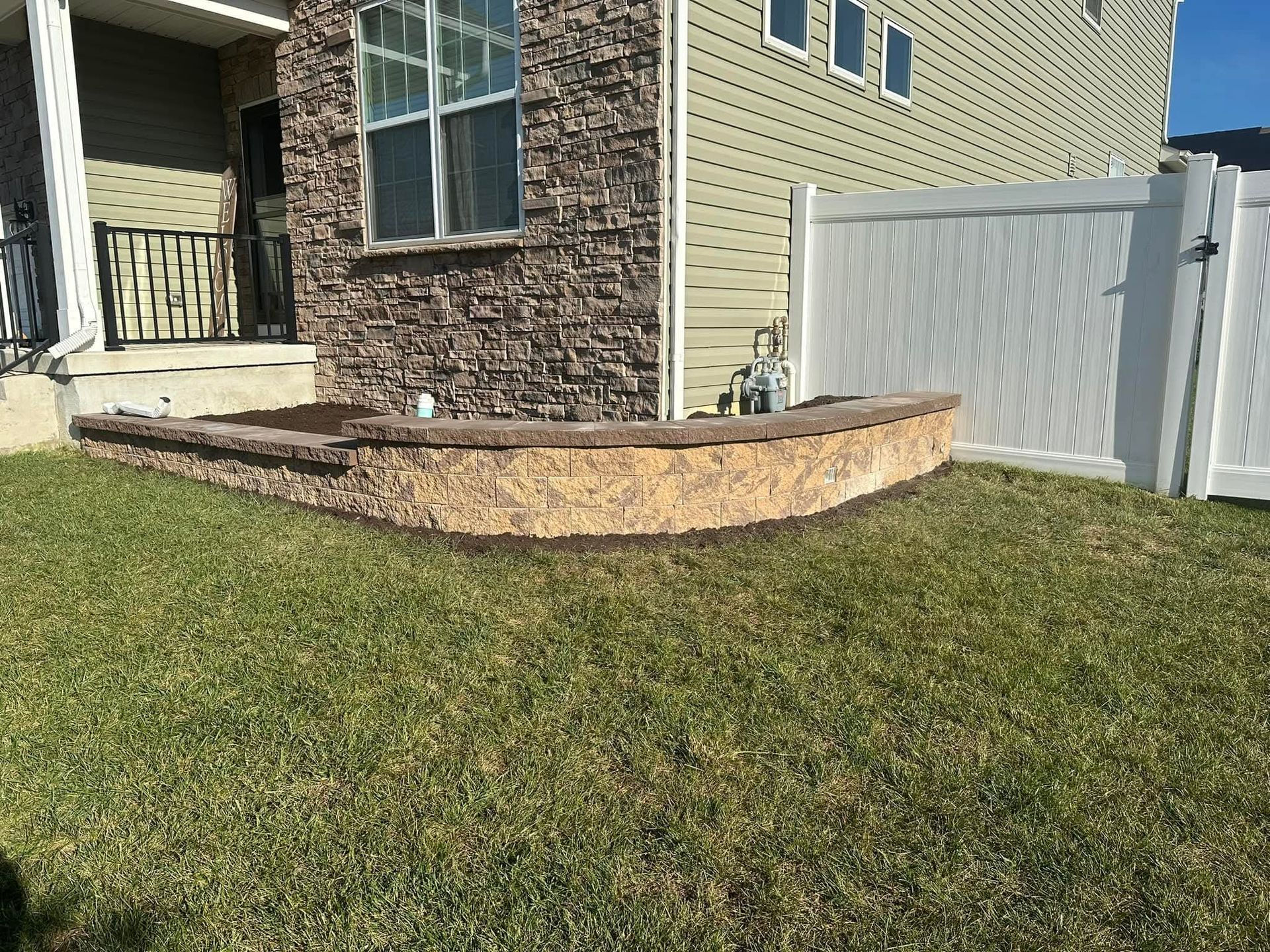 Tan stone retaining wall in front yard with green grass and white fence.