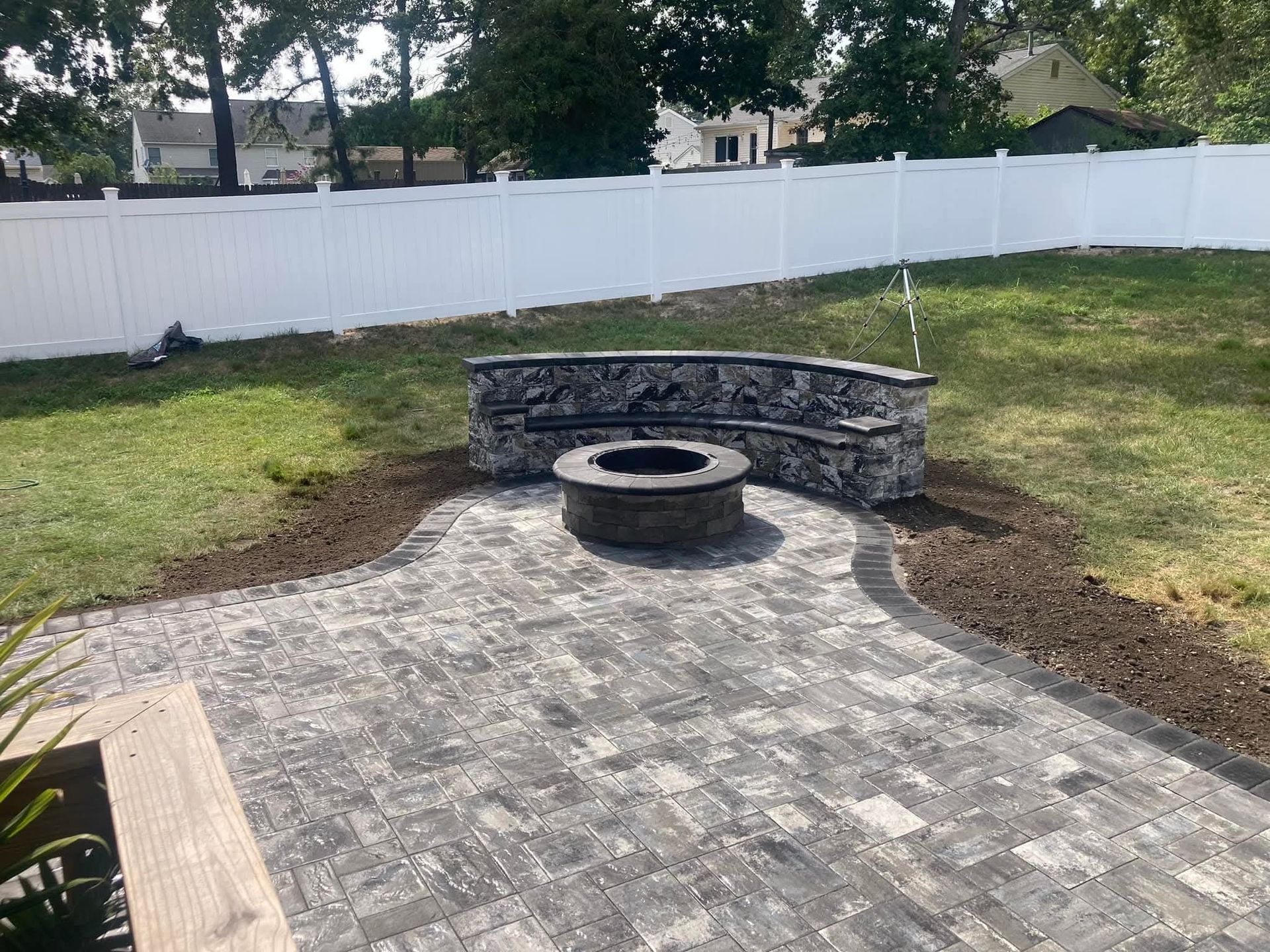 Stone patio with fire pit, built-in stone bench, and white fence in a backyard.