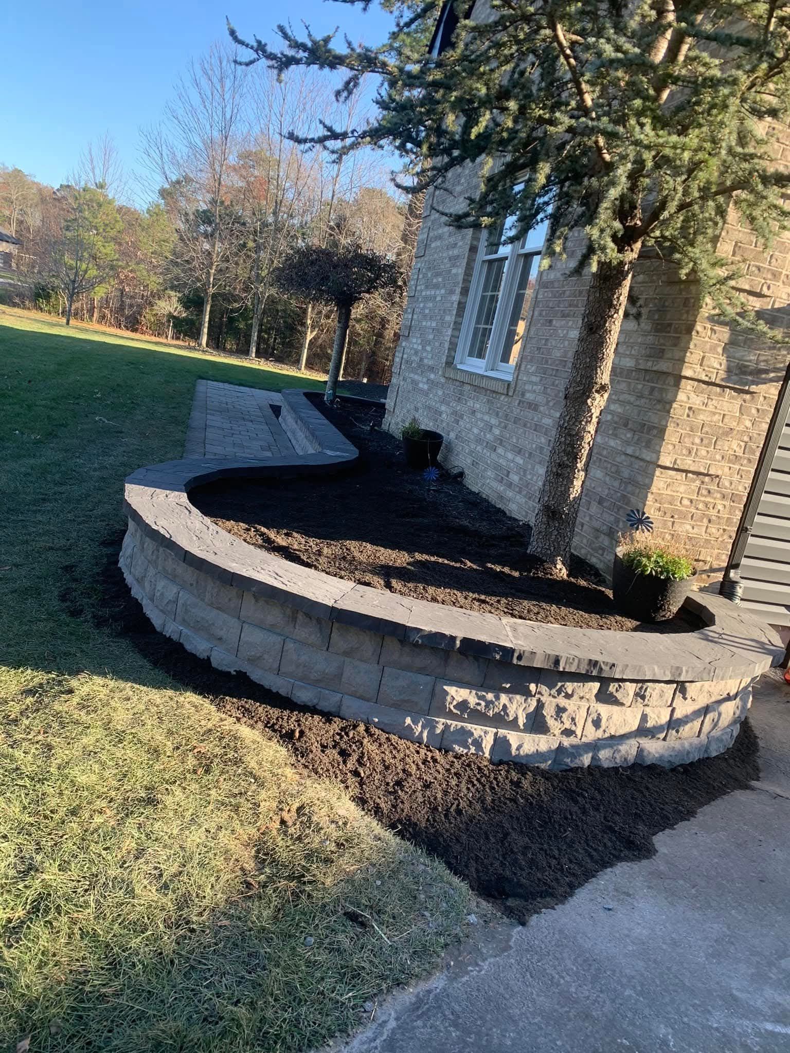 A curved retaining wall with dark mulch borders a house; a tree leans over it in daylight.