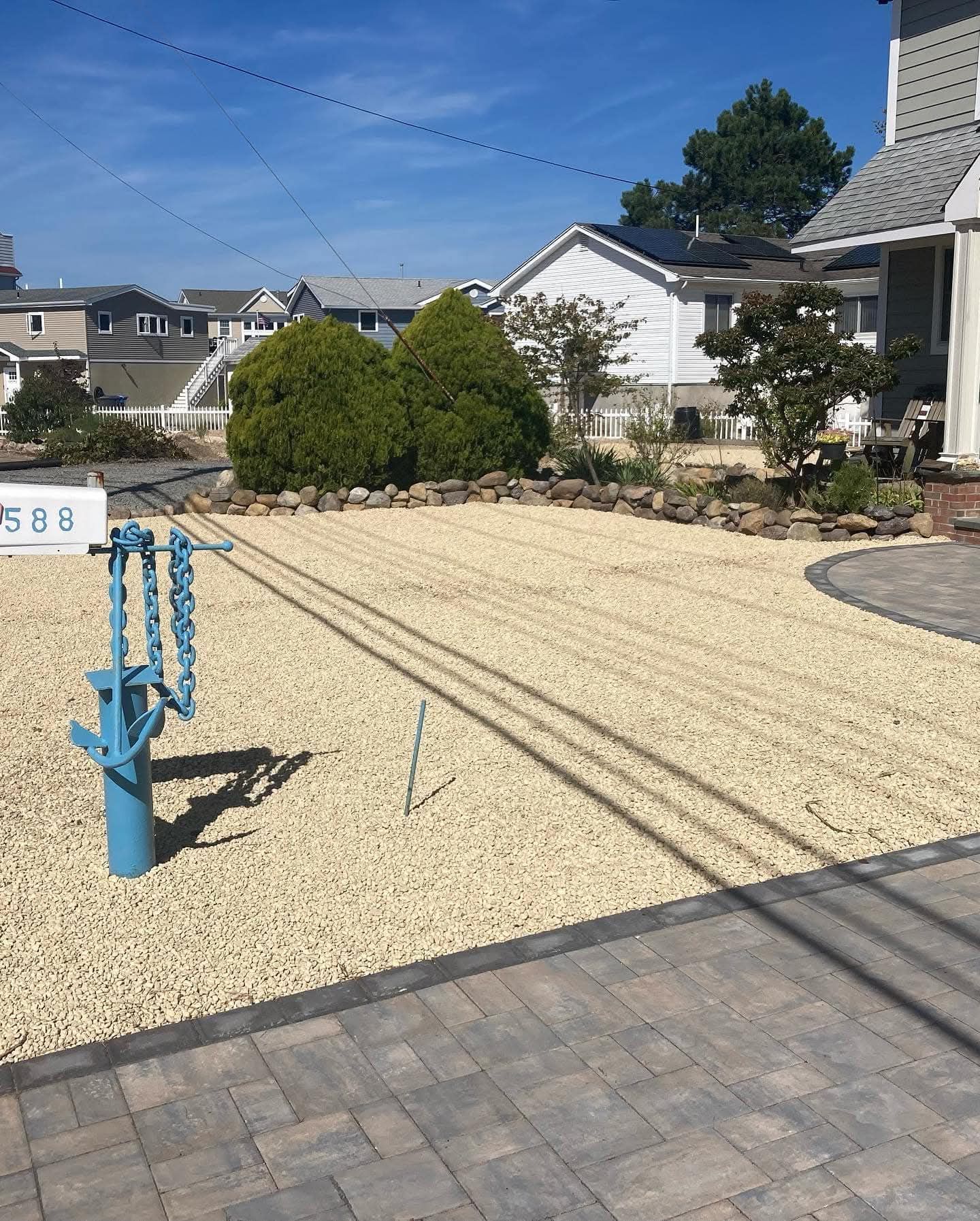 A gravel-covered front yard with a blue sculpture; houses in the background under a blue sky.