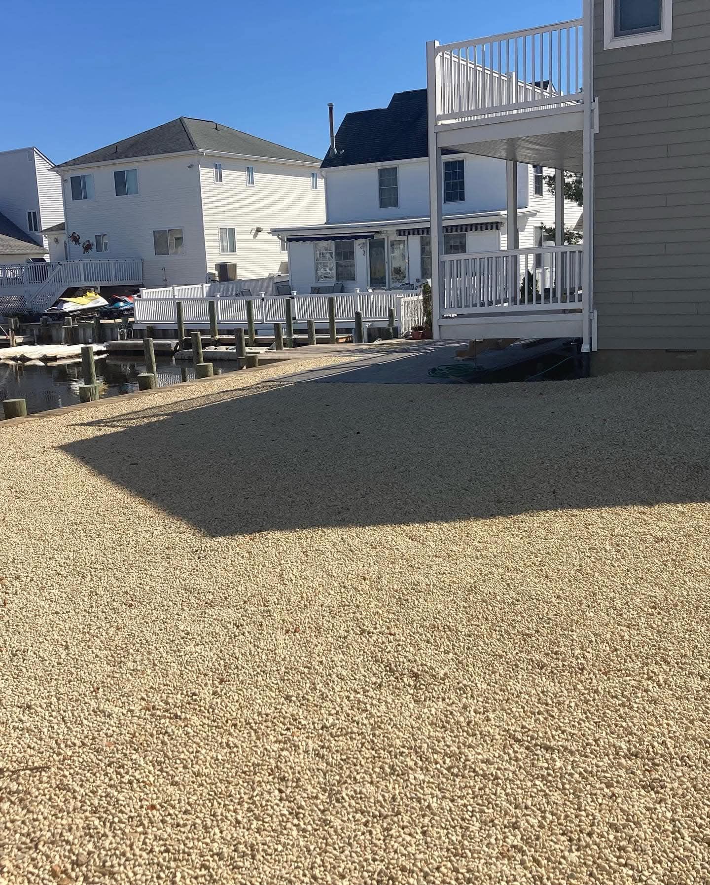 A gravel yard next to a light-colored building with a deck, overlooking a waterway with docks.