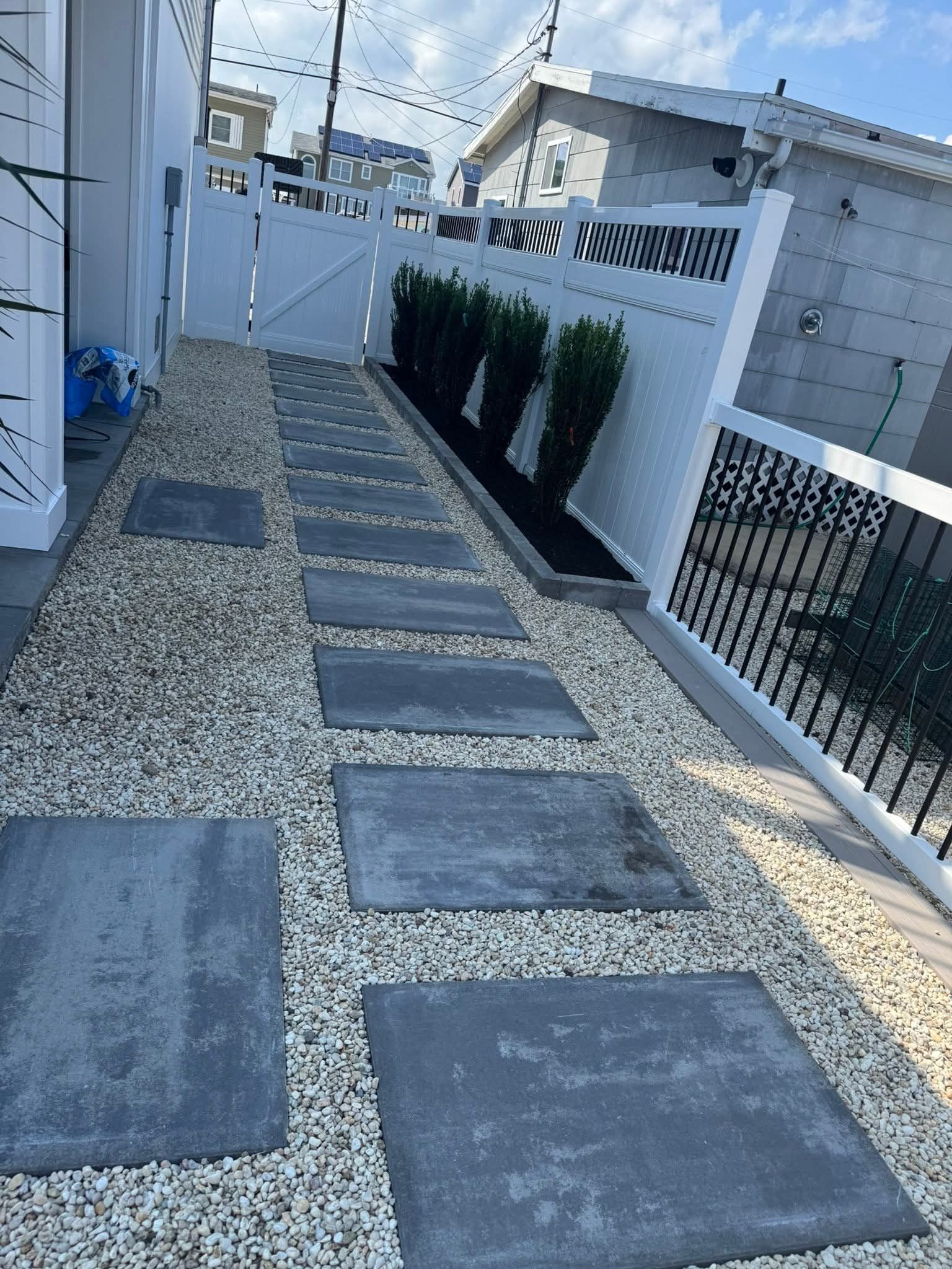 Stone pathway with stepping stones and gravel, bordered by a white fence and small green bushes.