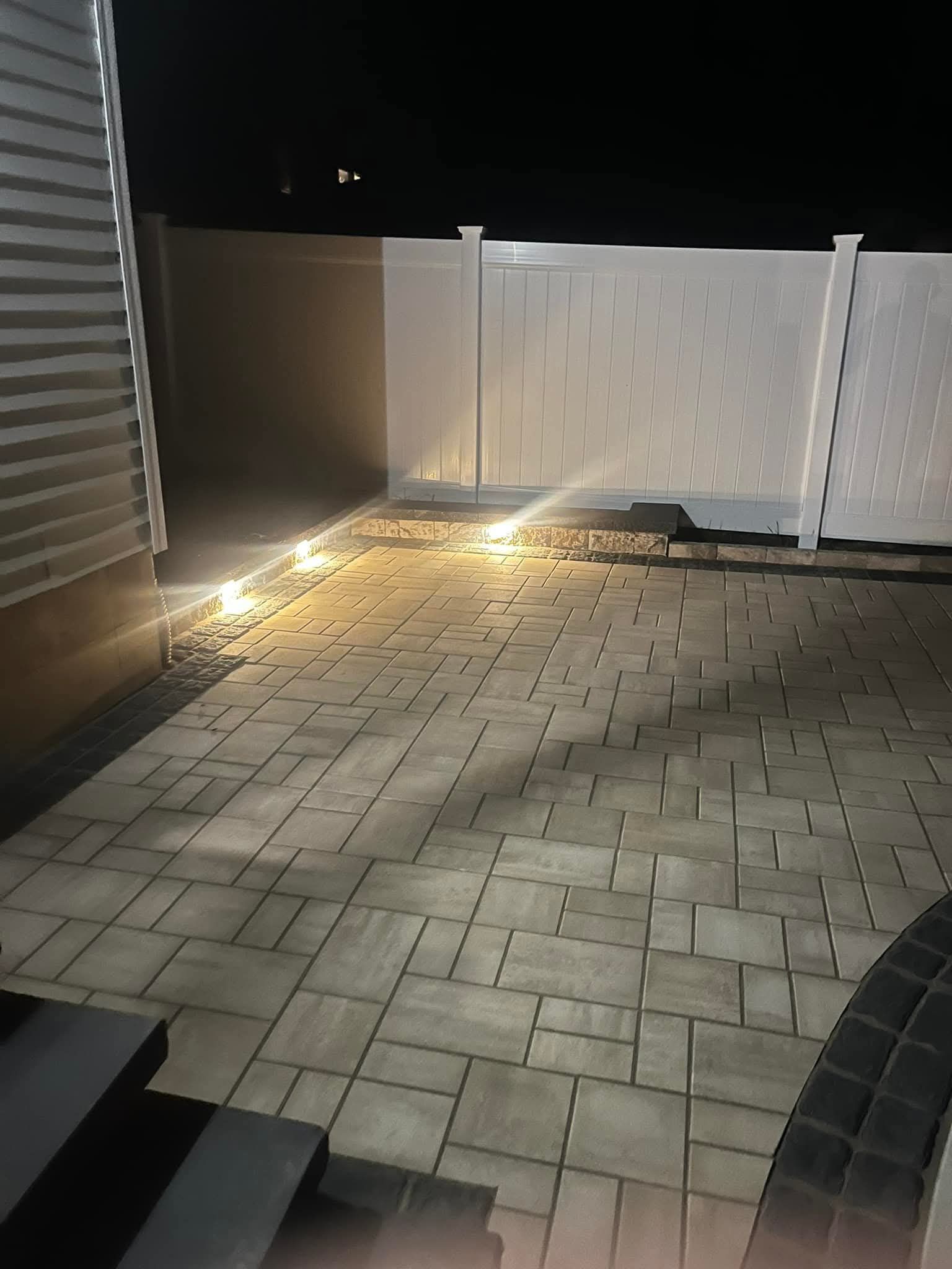 Nighttime outdoor patio with pavers, white fence, and string lights.