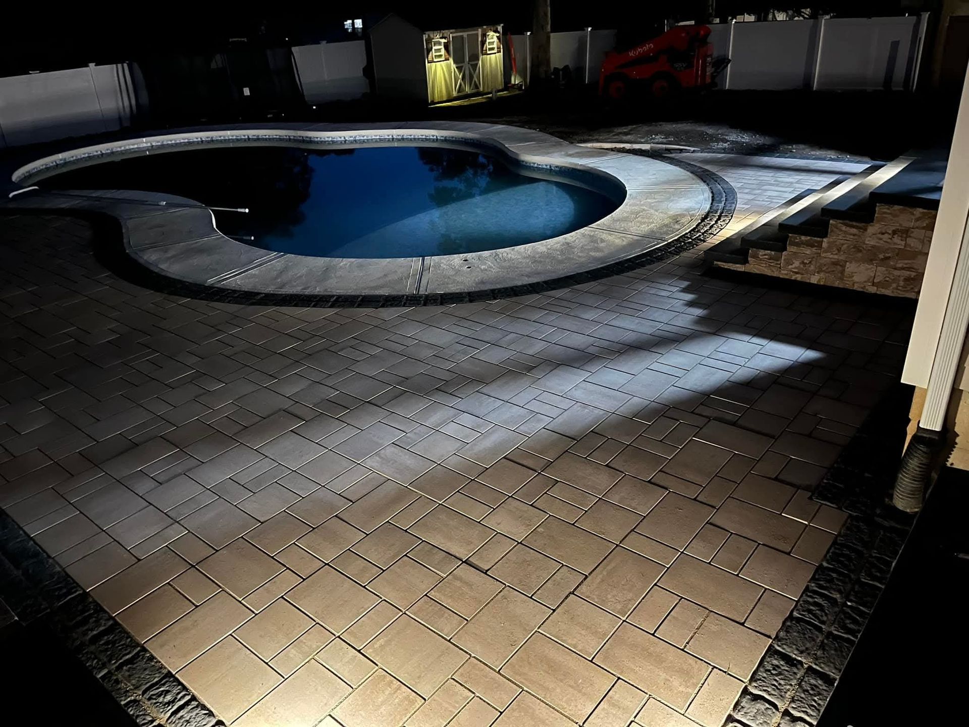 Nighttime backyard scene with a dark pool, paved patio, and ambient lighting.