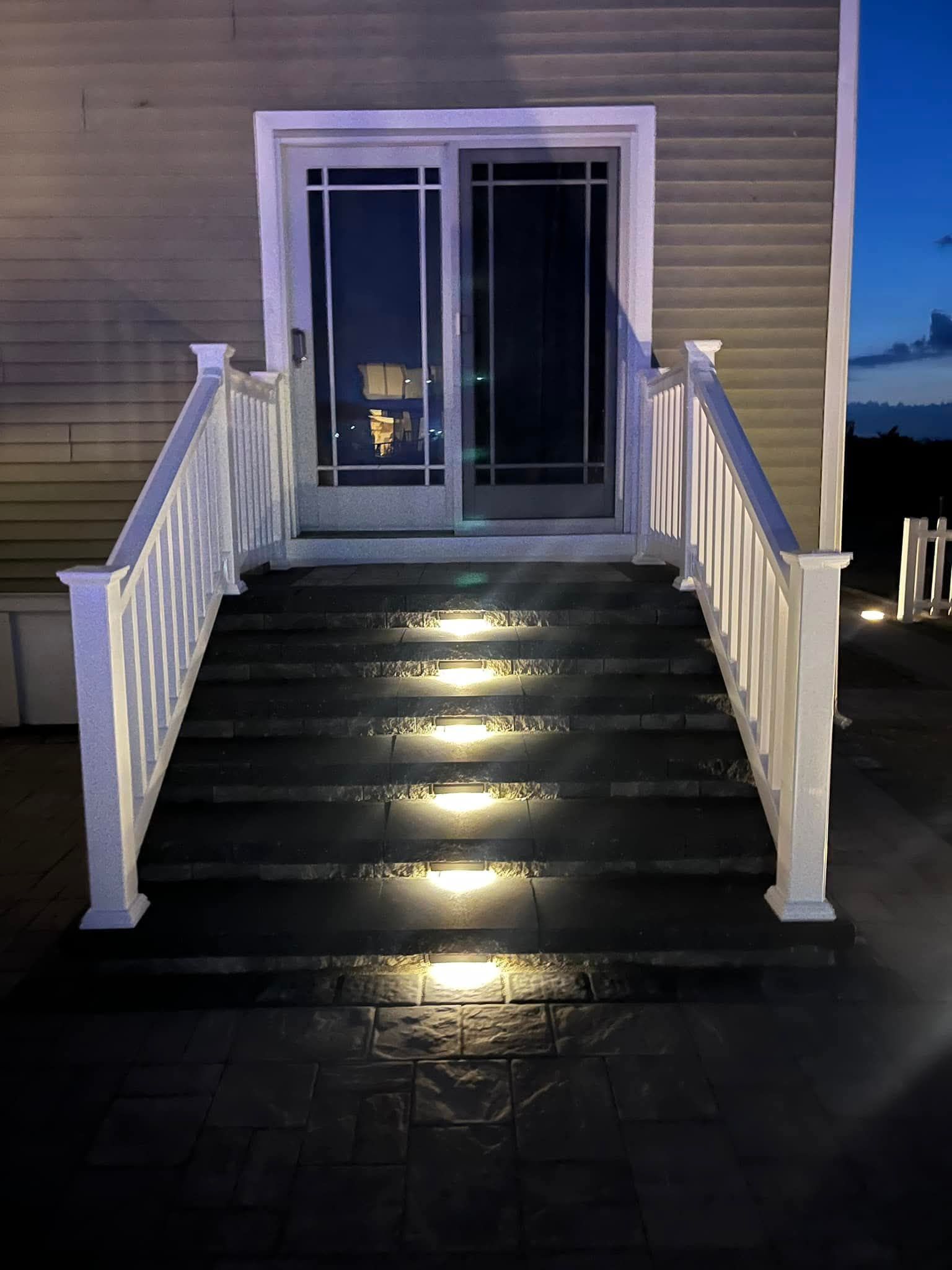 White-railed steps with built-in lights lead to a sliding glass door. Dark exterior at dusk.