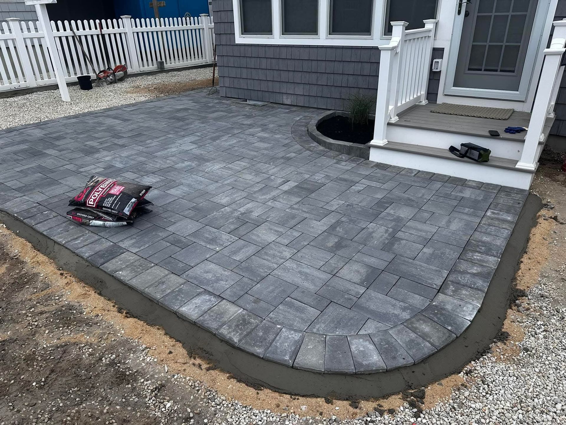A gray paver patio with curved edges, leading to a house entrance, with a bag of pavers in the foreground.